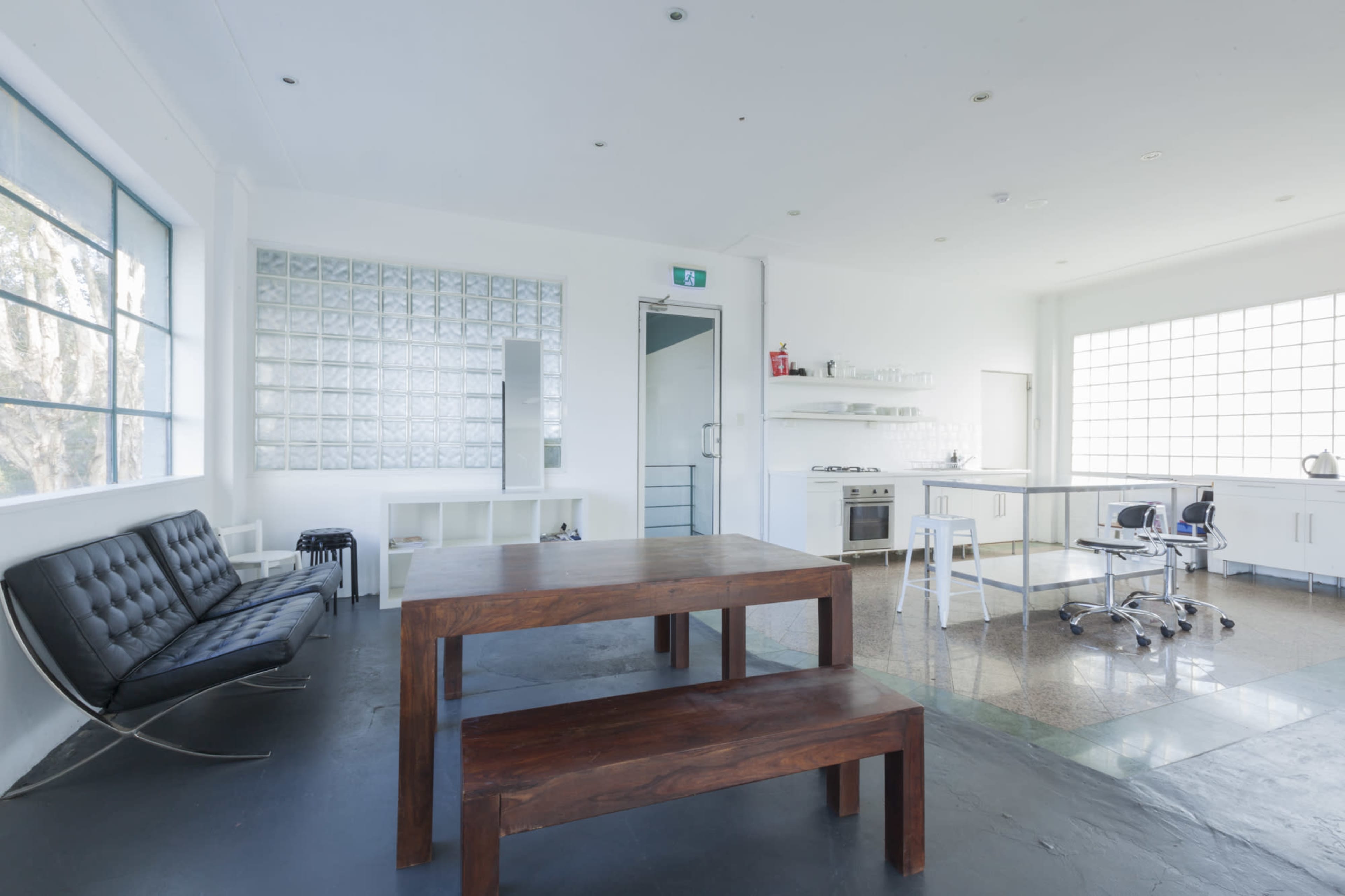 A modern, minimalist kitchen and dining area with a wooden table, a black sofa, and large windows allowing natural light.