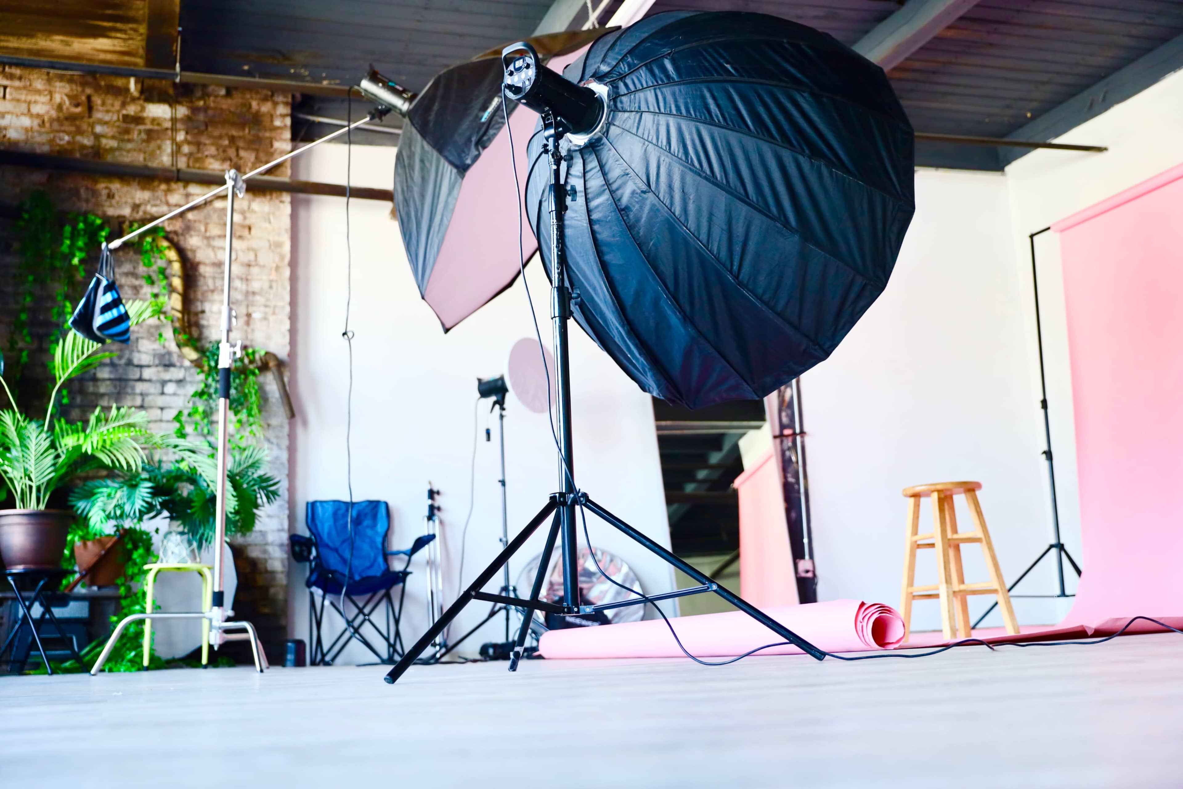 A photography studio with lighting equipment, a wooden stool, and a pink backdrop rolled on the floor.