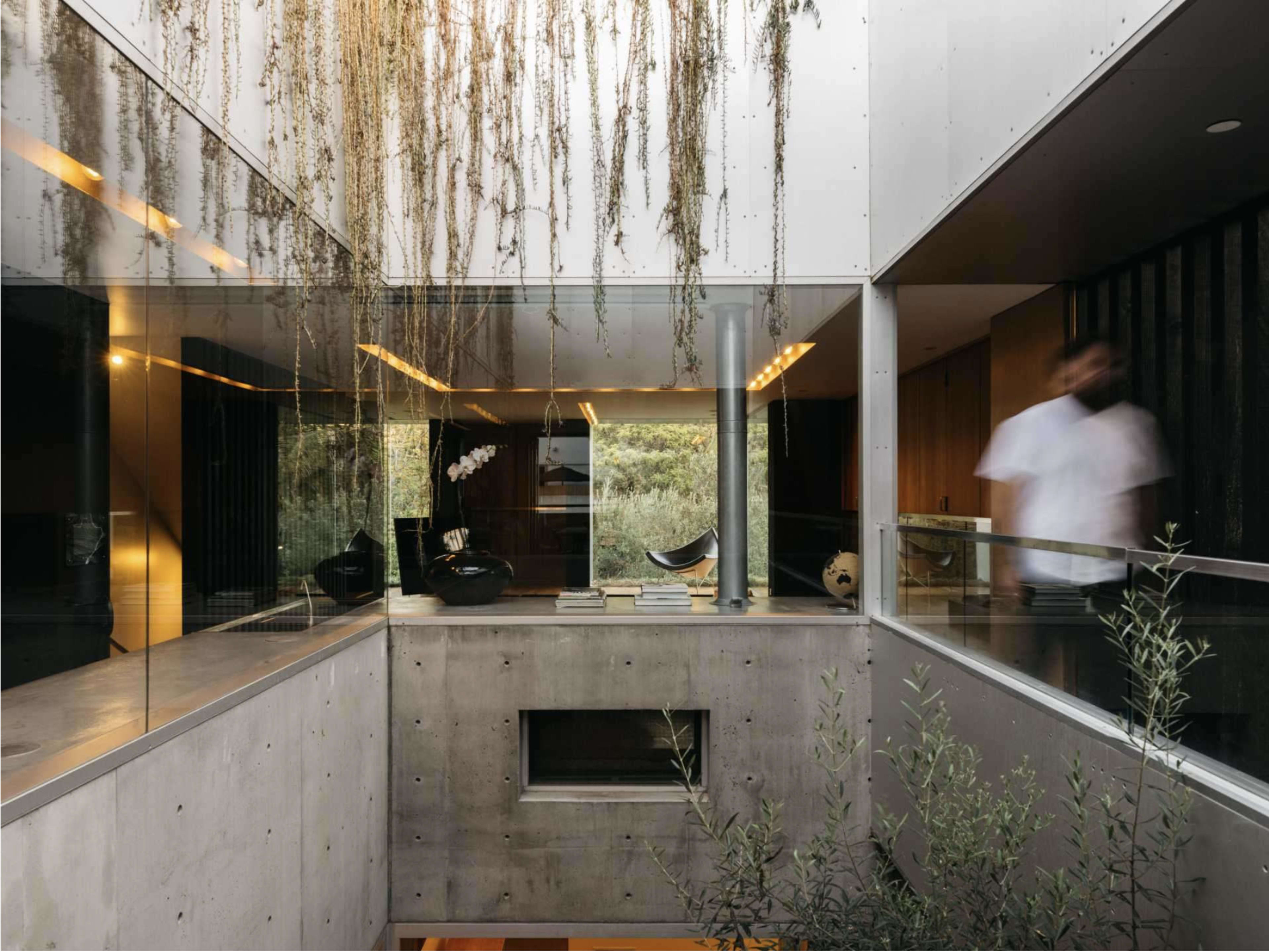 A modern interior with concrete and glass elements, featuring a central atrium with hanging foliage and a person walking through the space.