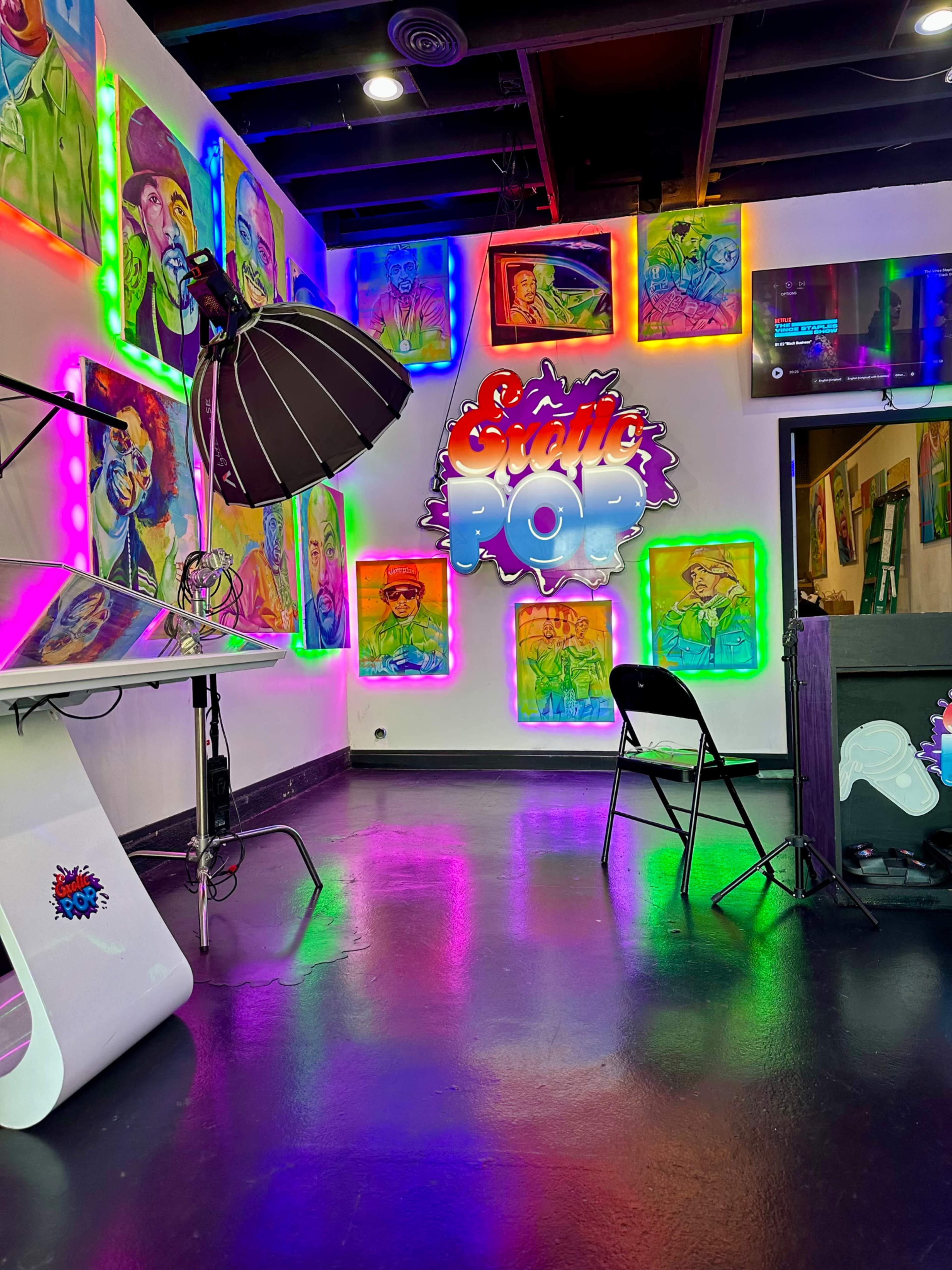 The image shows a brightly lit art studio with colorful portraits hanging on the walls and a sign that reads "Sattle Pop" illuminated in vibrant colors.