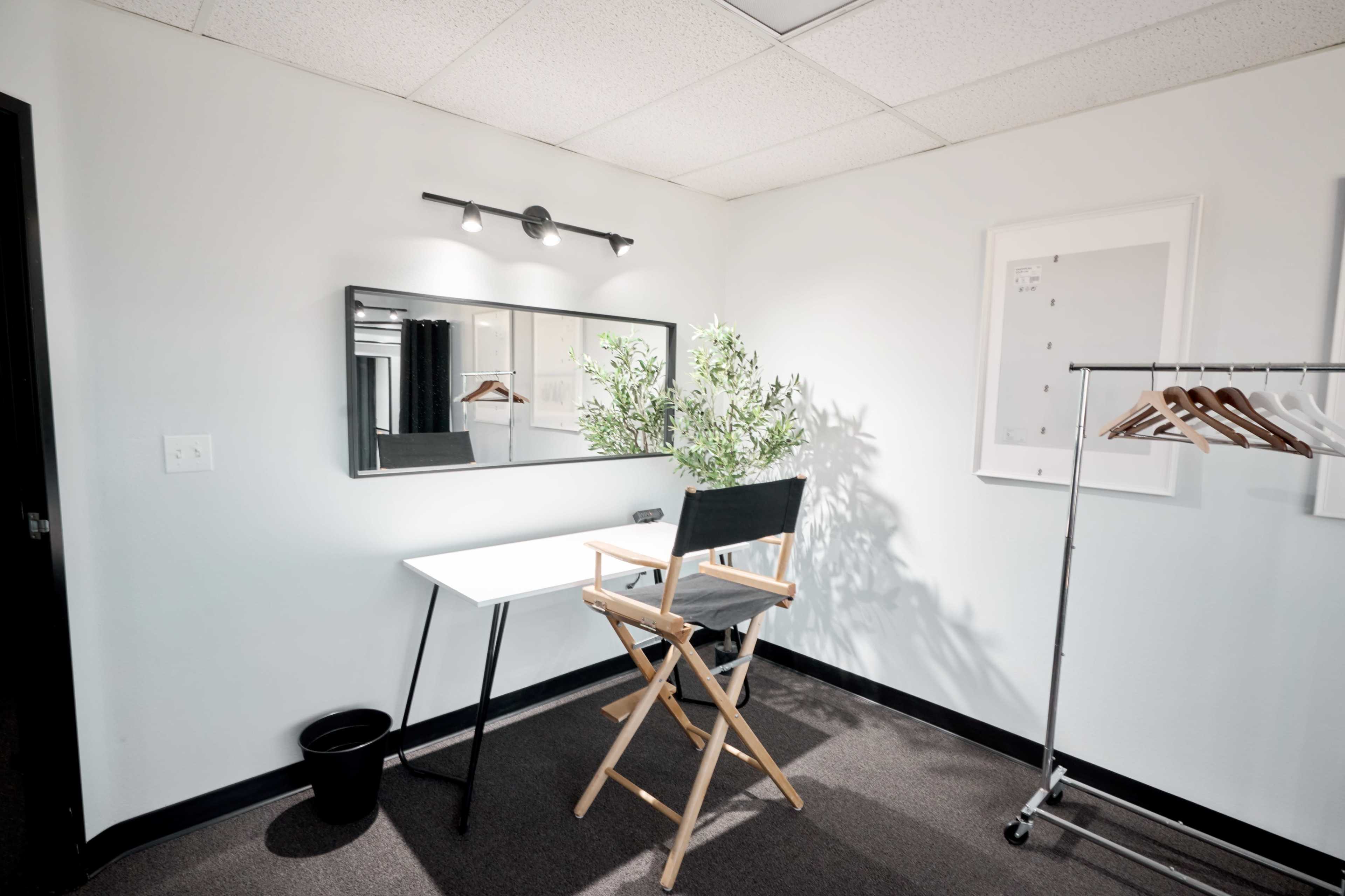 A small room with a chair, a mirror, a table, a potted plant, and a clothing rack with hangers.