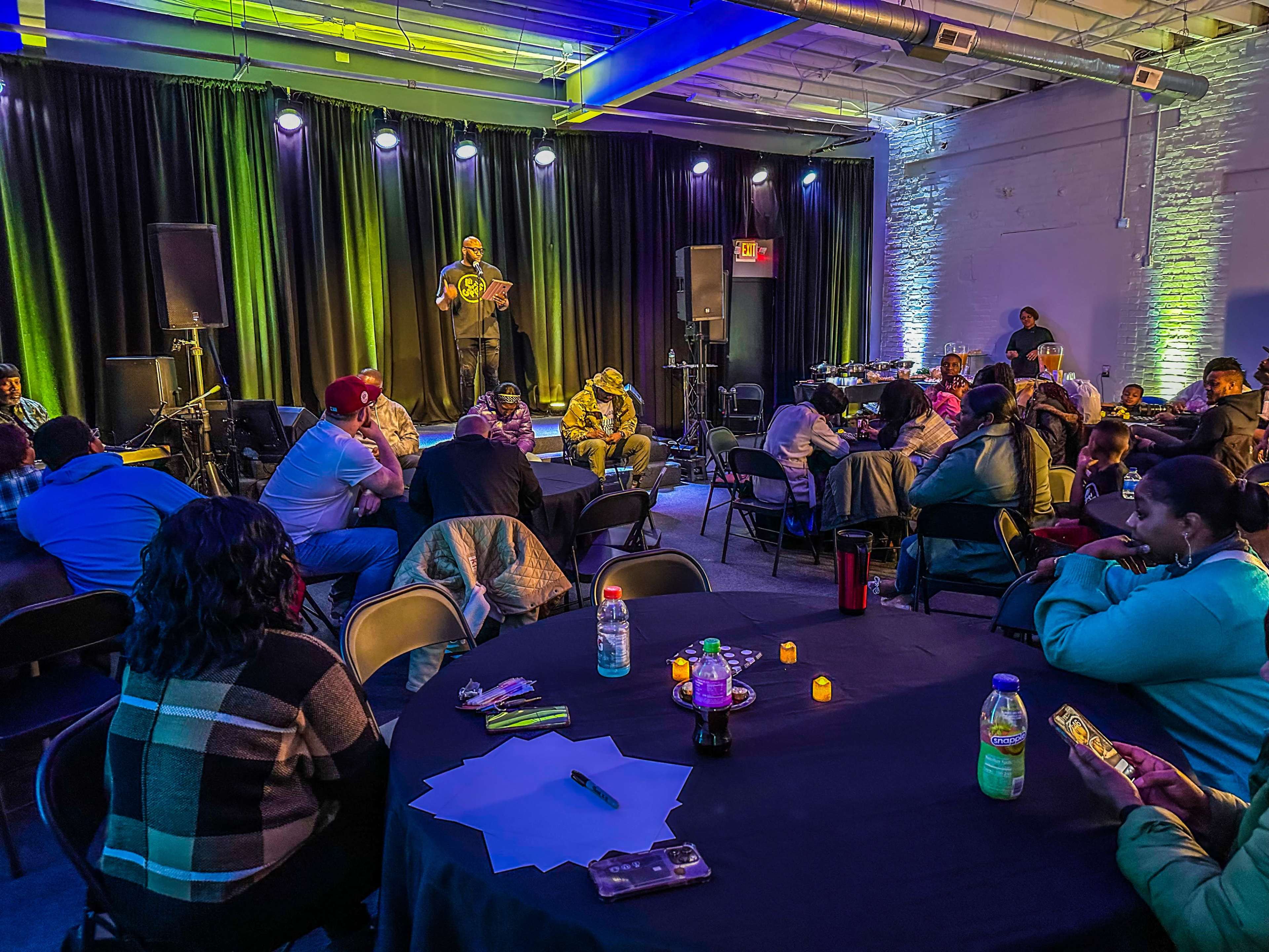 A performer stands on stage reading in front of an audience seated at round tables in a dimly lit venue.