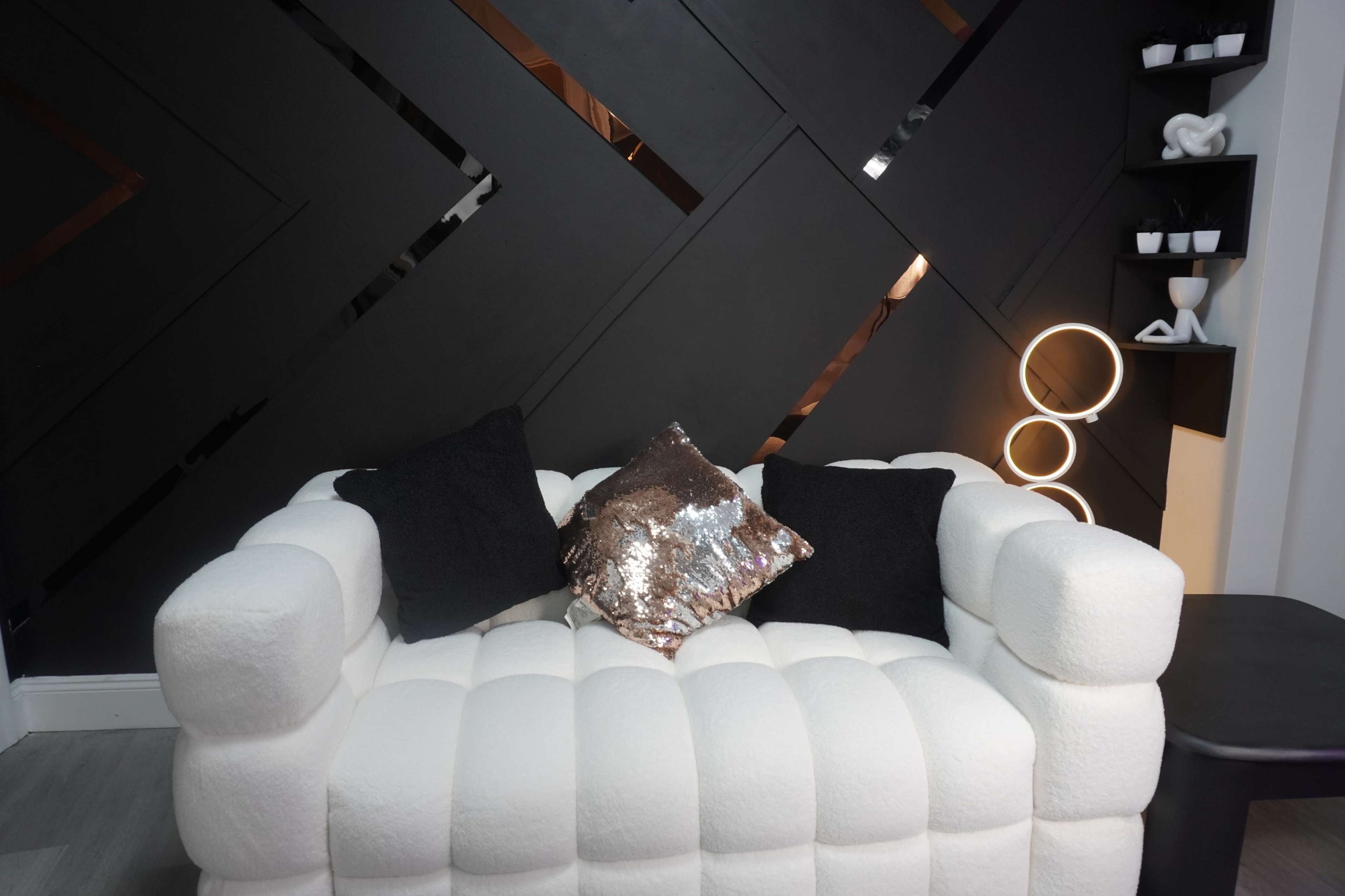 A white tufted couch adorned with black and sequin throw pillows, set against a geometric black wall with a minimalist decor piece nearby.