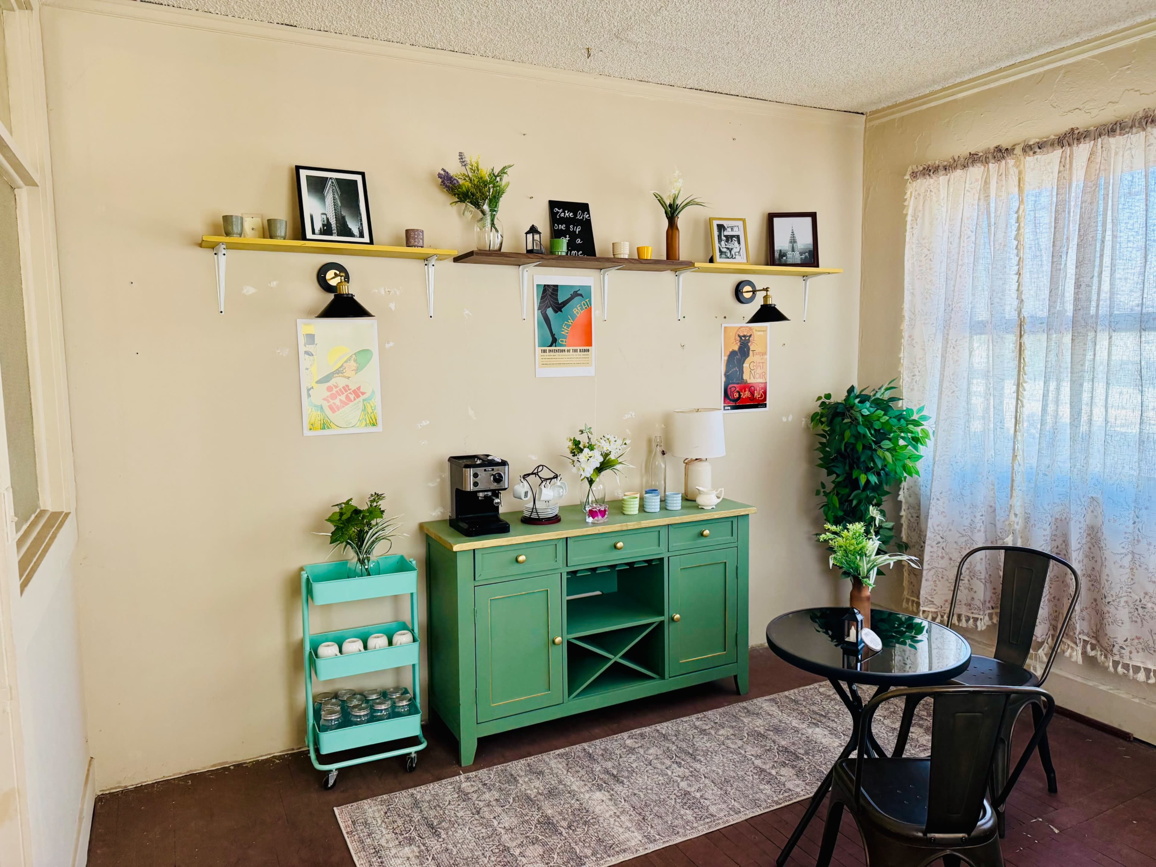 The image shows a bright, cozy room featuring a green sideboard, a small round table with chairs, and decorative shelves displaying plants and artwork.