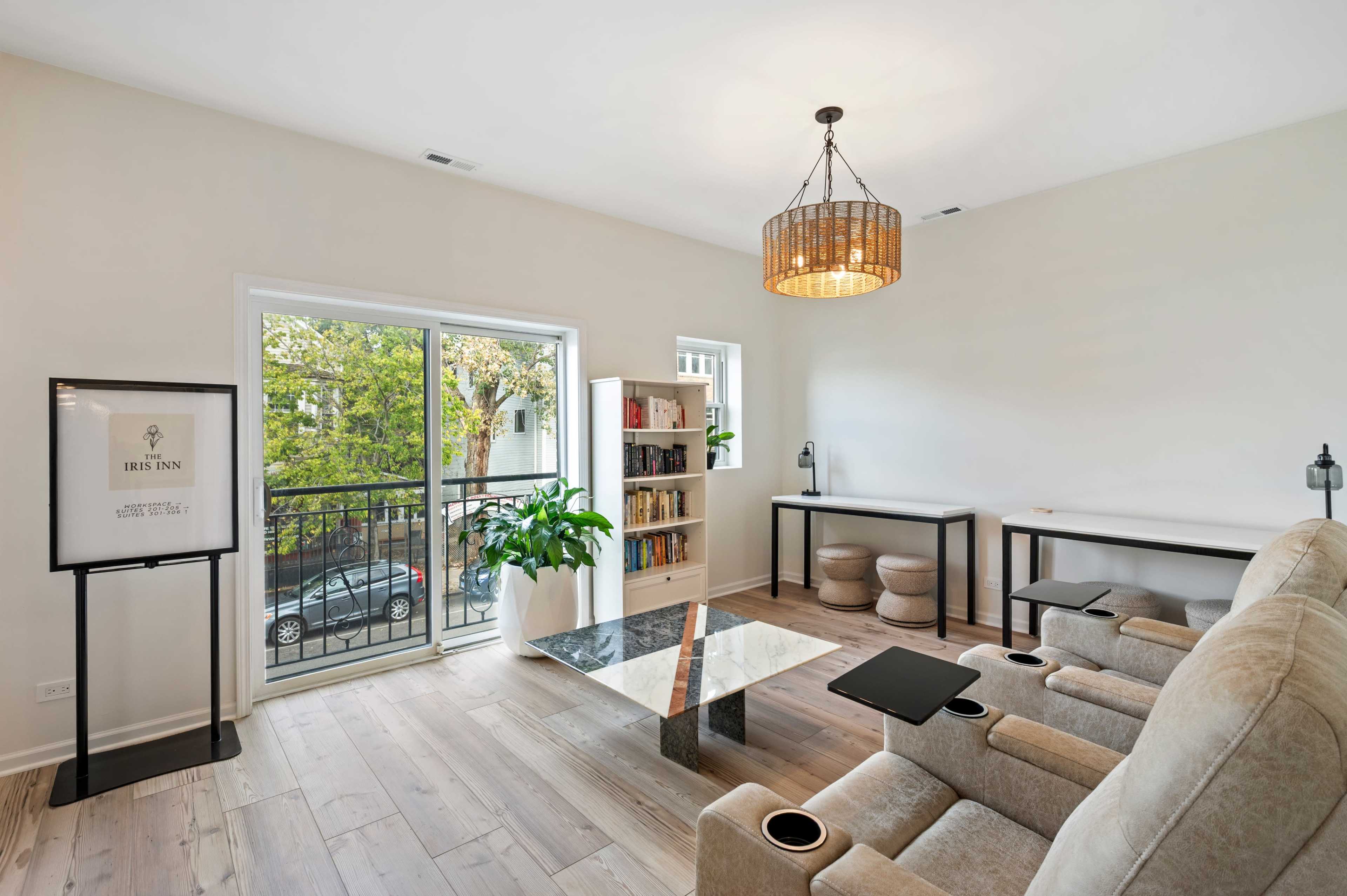 The image shows a modern living room with two reclining chairs, a coffee table, shelves filled with books, and large windows overlooking a balcony.