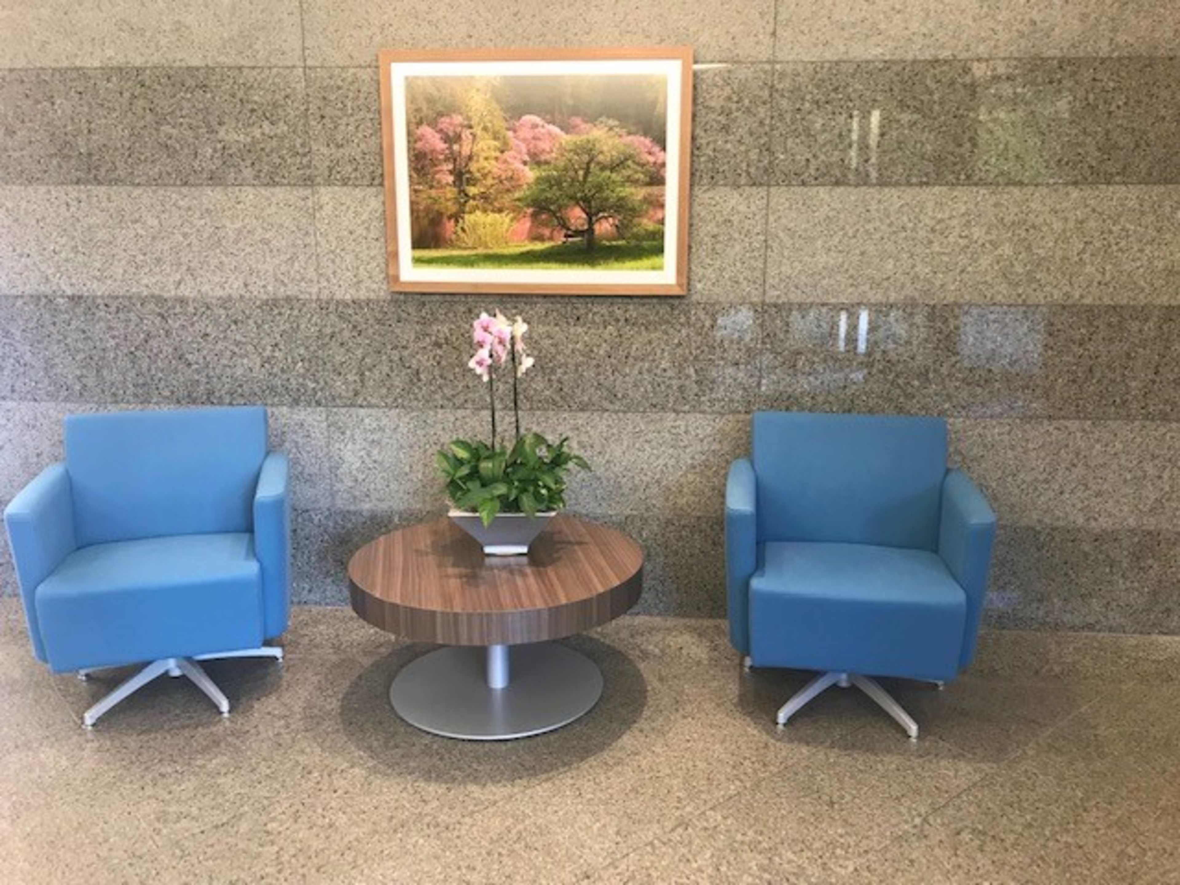 Two blue chairs flanking a circular wooden table with a potted orchid, set against a gray stone wall featuring a framed landscape photograph.