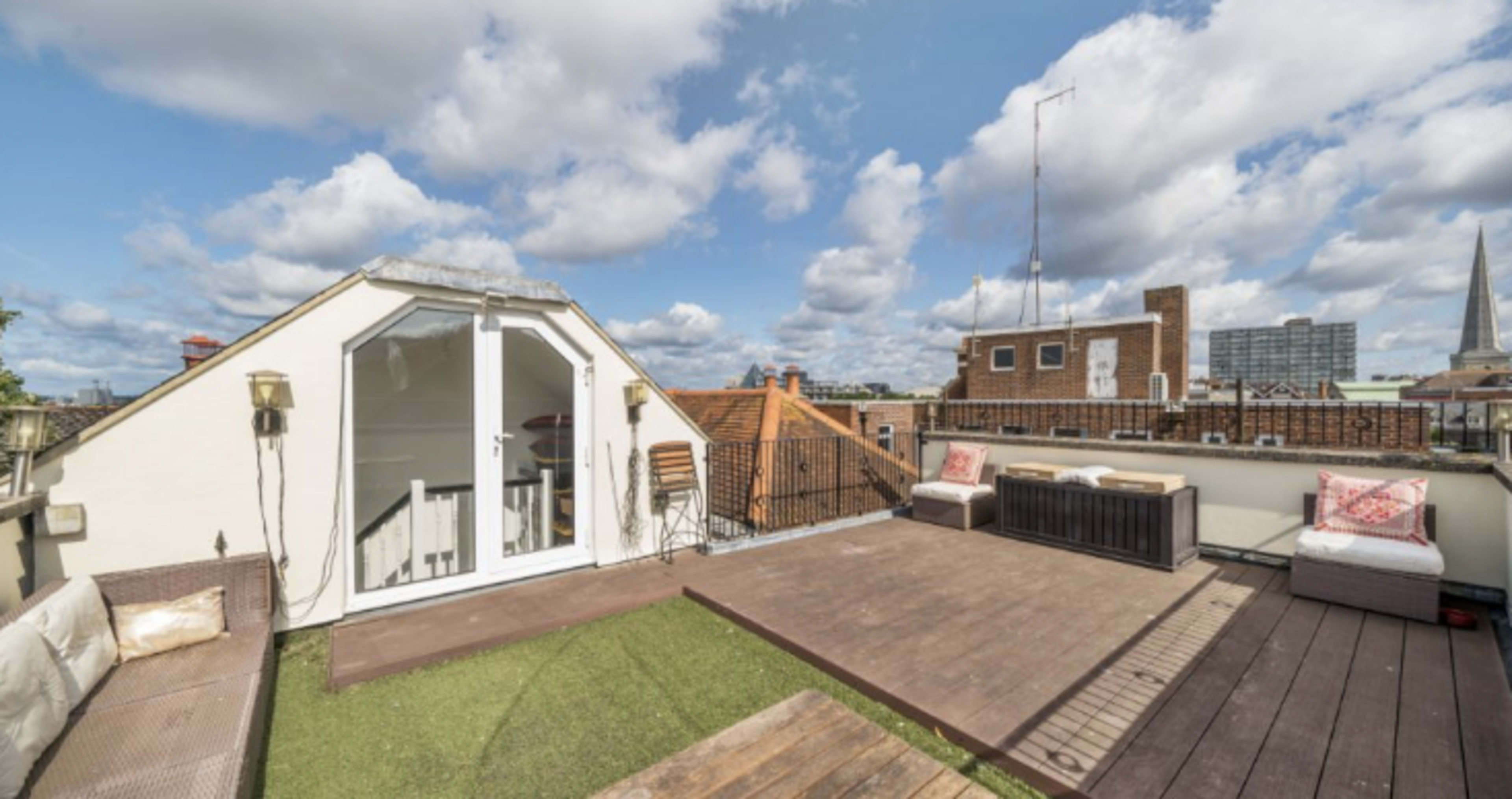 A rooftop terrace with seating areas, a wooden deck, and a view of surrounding buildings under a partly cloudy sky.