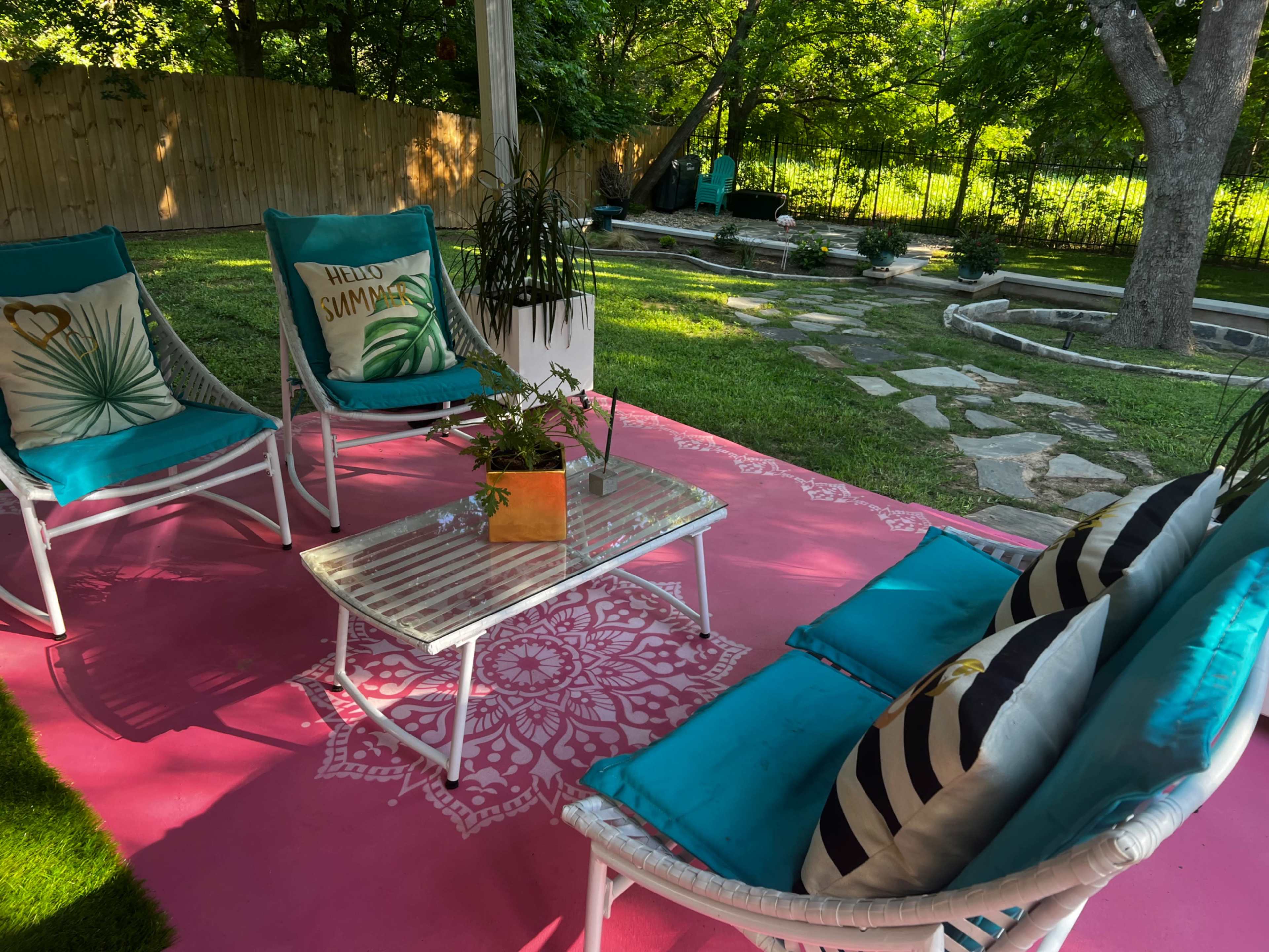 A backyard seating area features white wicker chairs with blue cushions and a pink patterned rug, surrounded by greenery and stone pathways.