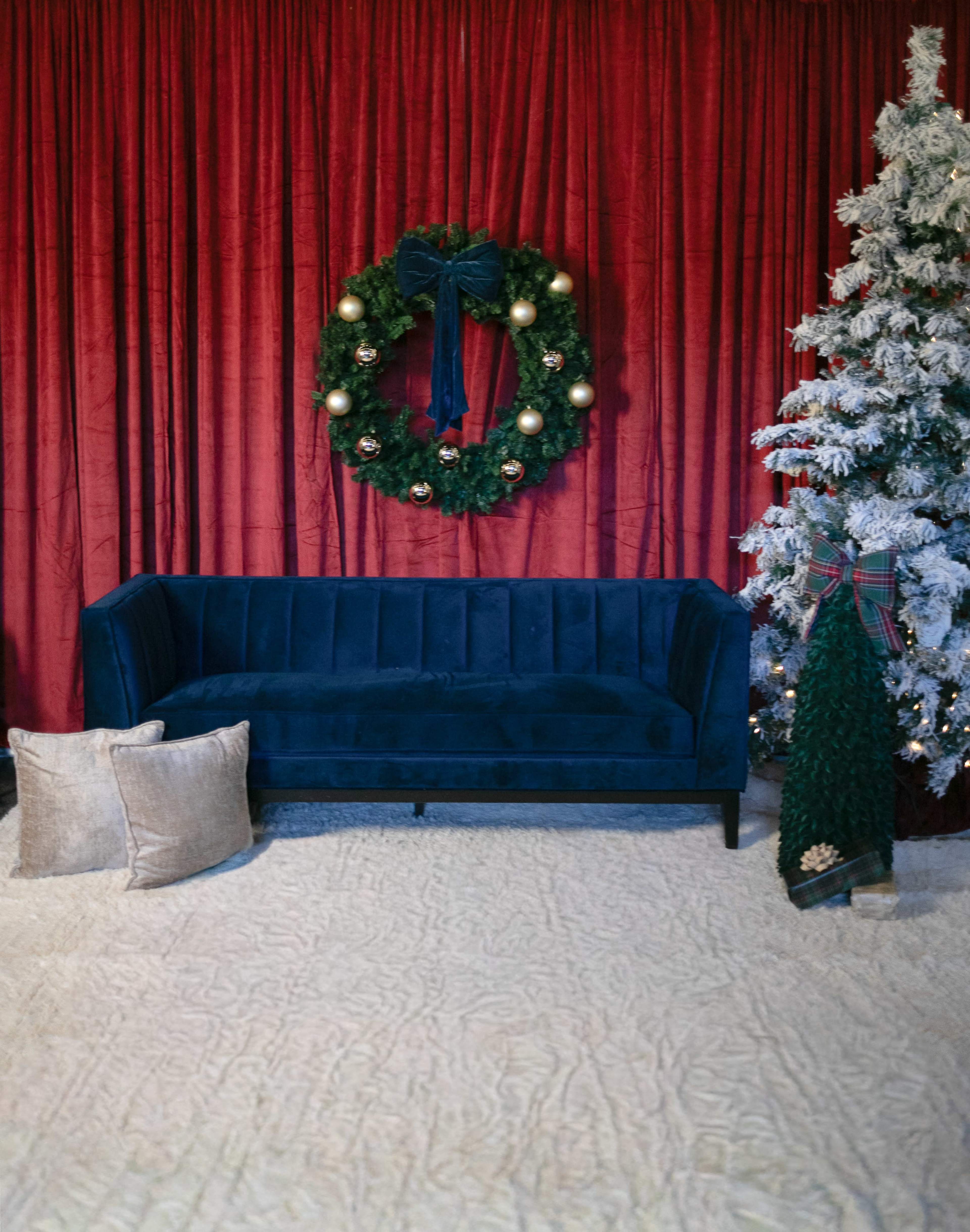 A blue sofa is positioned in front of a red curtain, with a decorated white Christmas tree and a wreath hanging on the wall.