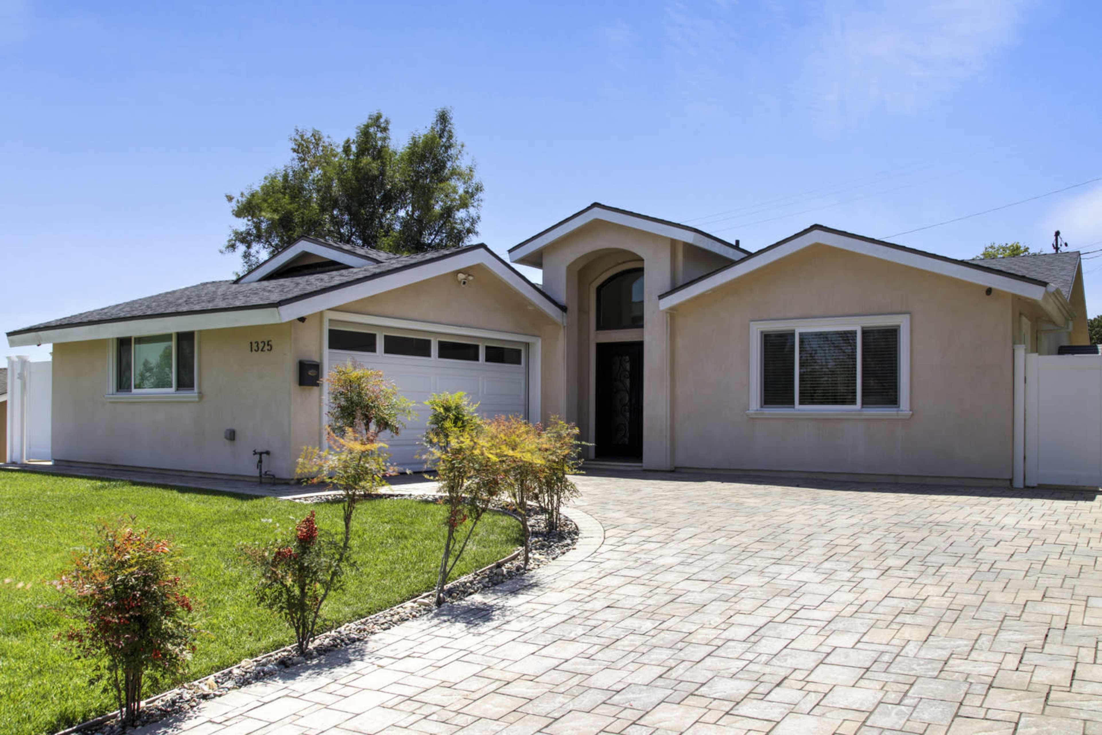 The image shows a single-story residential home with a paved driveway, manicured front lawn, and a combination of a garage and entrance door.