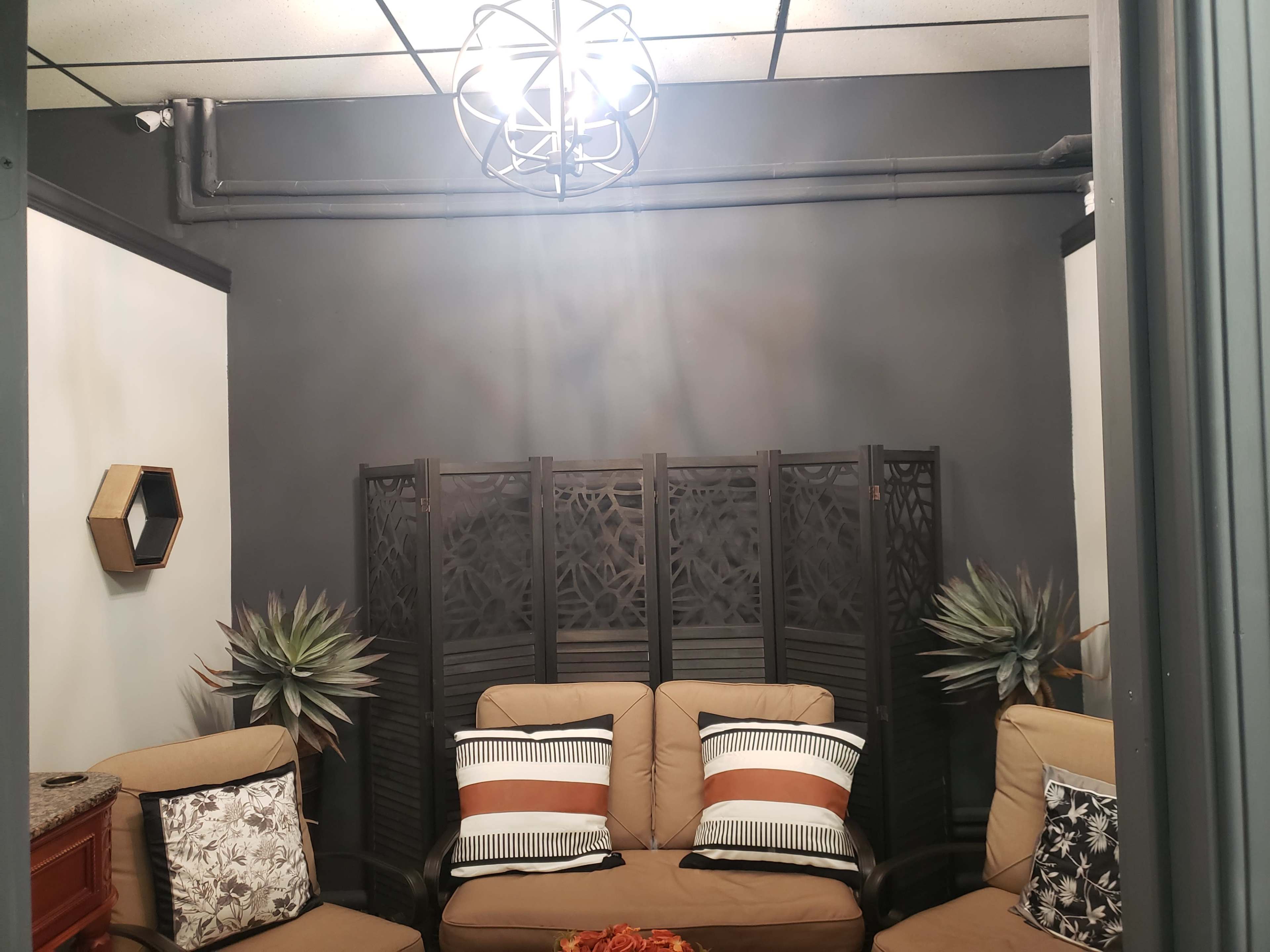 The image shows a small, modern lounge area with a three-seat sofa, decorative cushions, a folding wooden screen, and artificial plants.