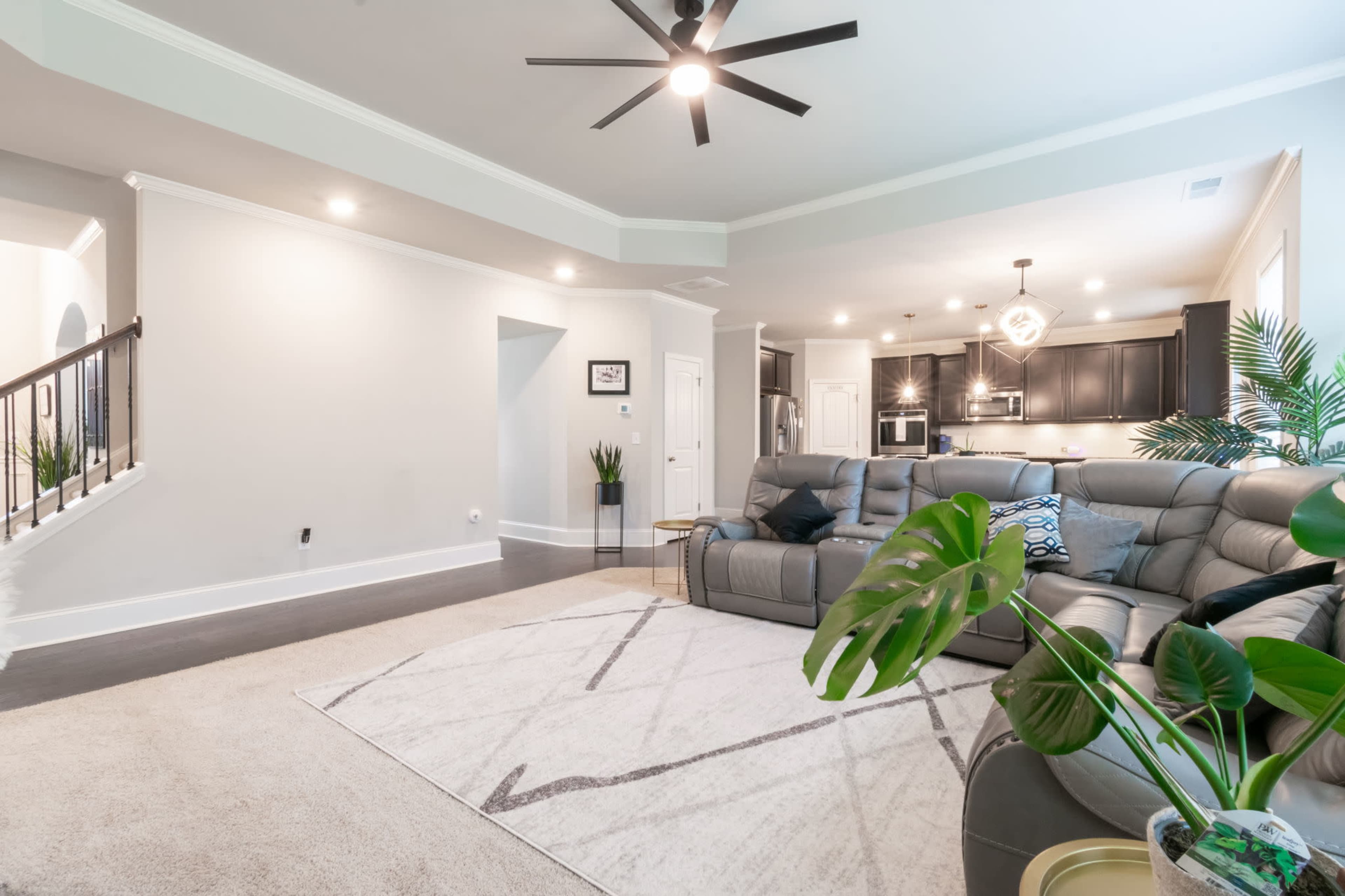 A spacious living room with a sectional sofa, a large rug, and a staircase leading to another level, along with modern kitchen appliances visible in the background.