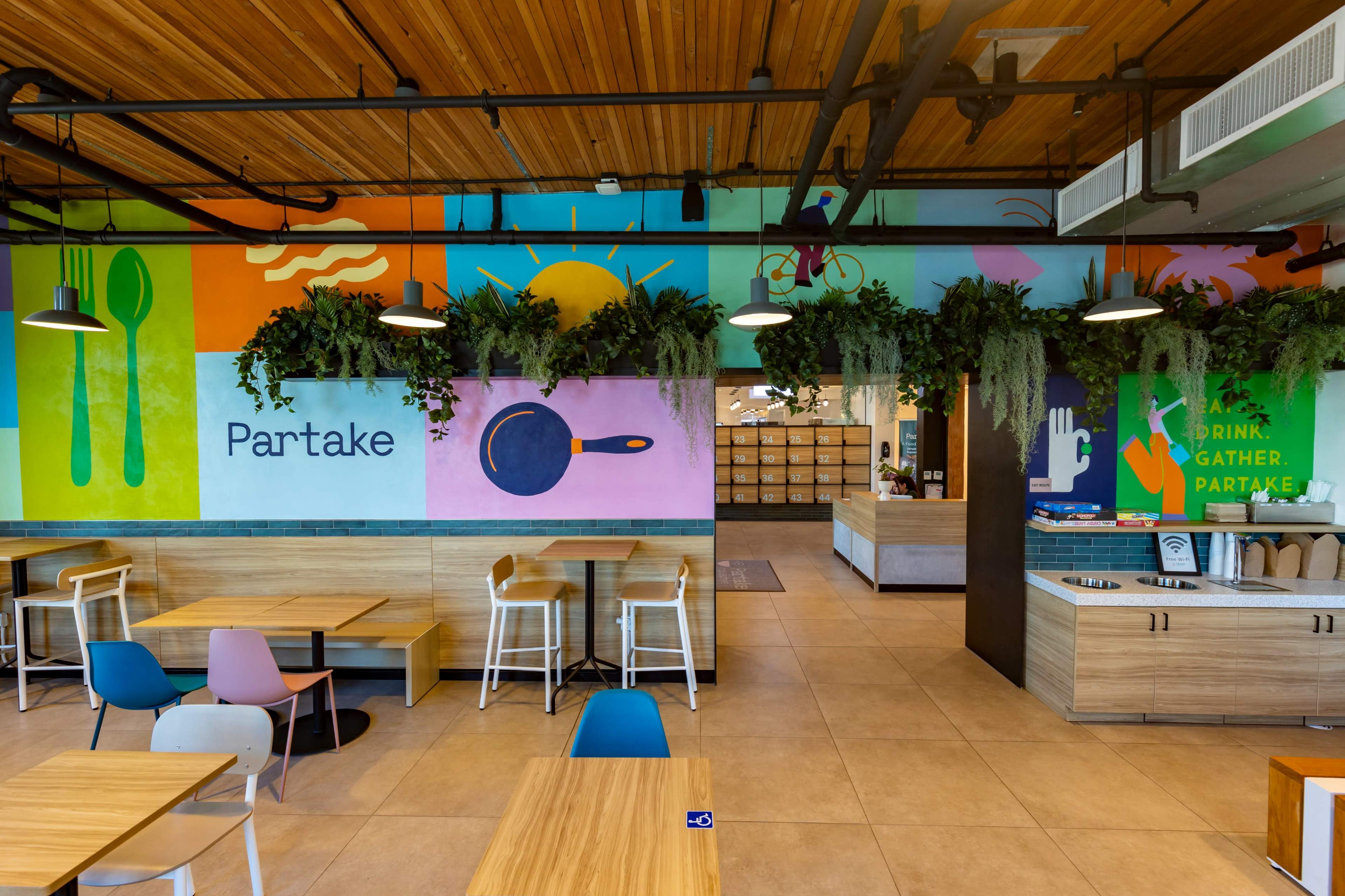 The image shows a colorful café interior with wooden tables, chairs, and vibrant wall murals featuring the word "Partake" and various graphic designs.