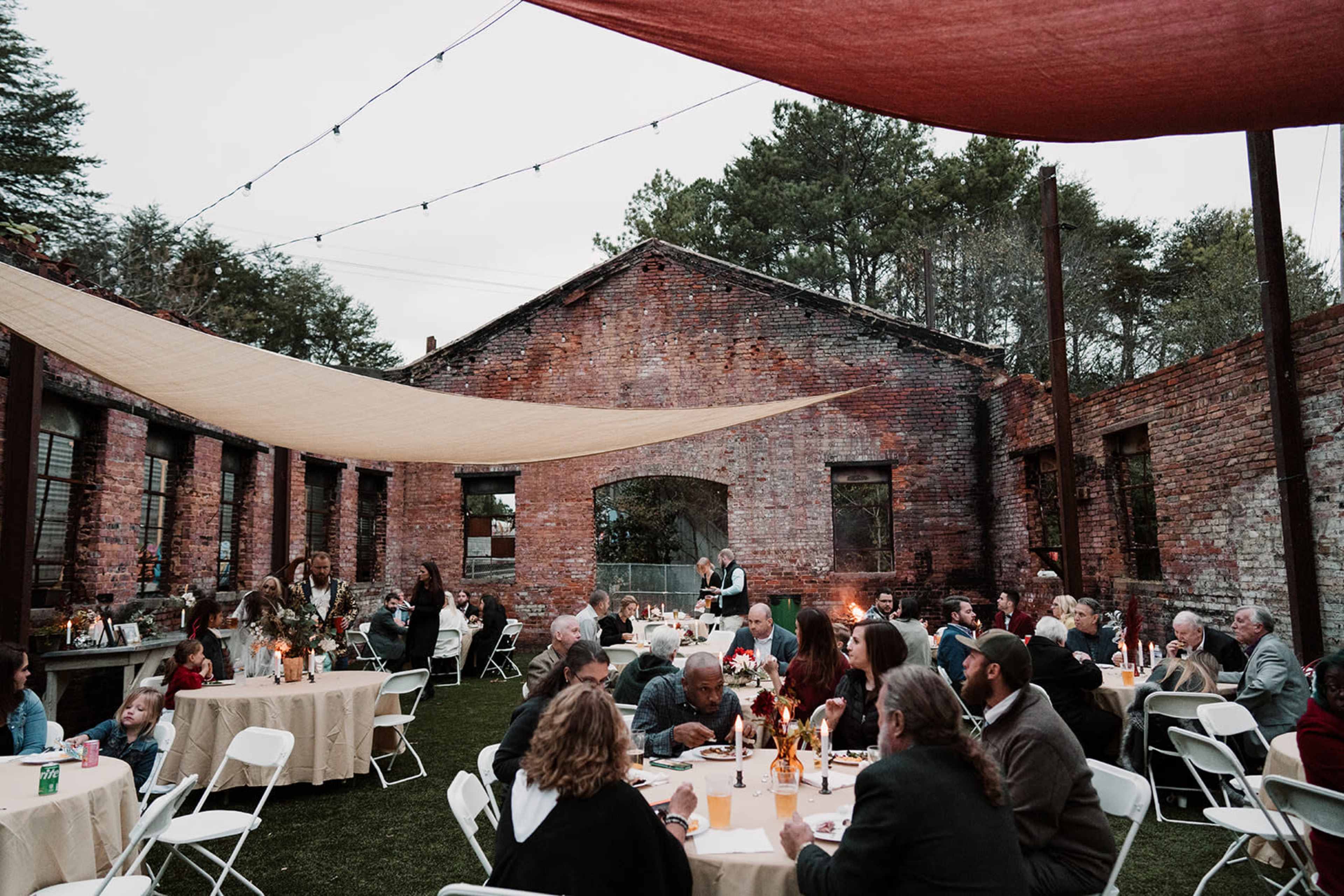 The scene depicts a gathering in an outdoor space with tables set up under a canopy, surrounded by a rustic brick building and greenery.