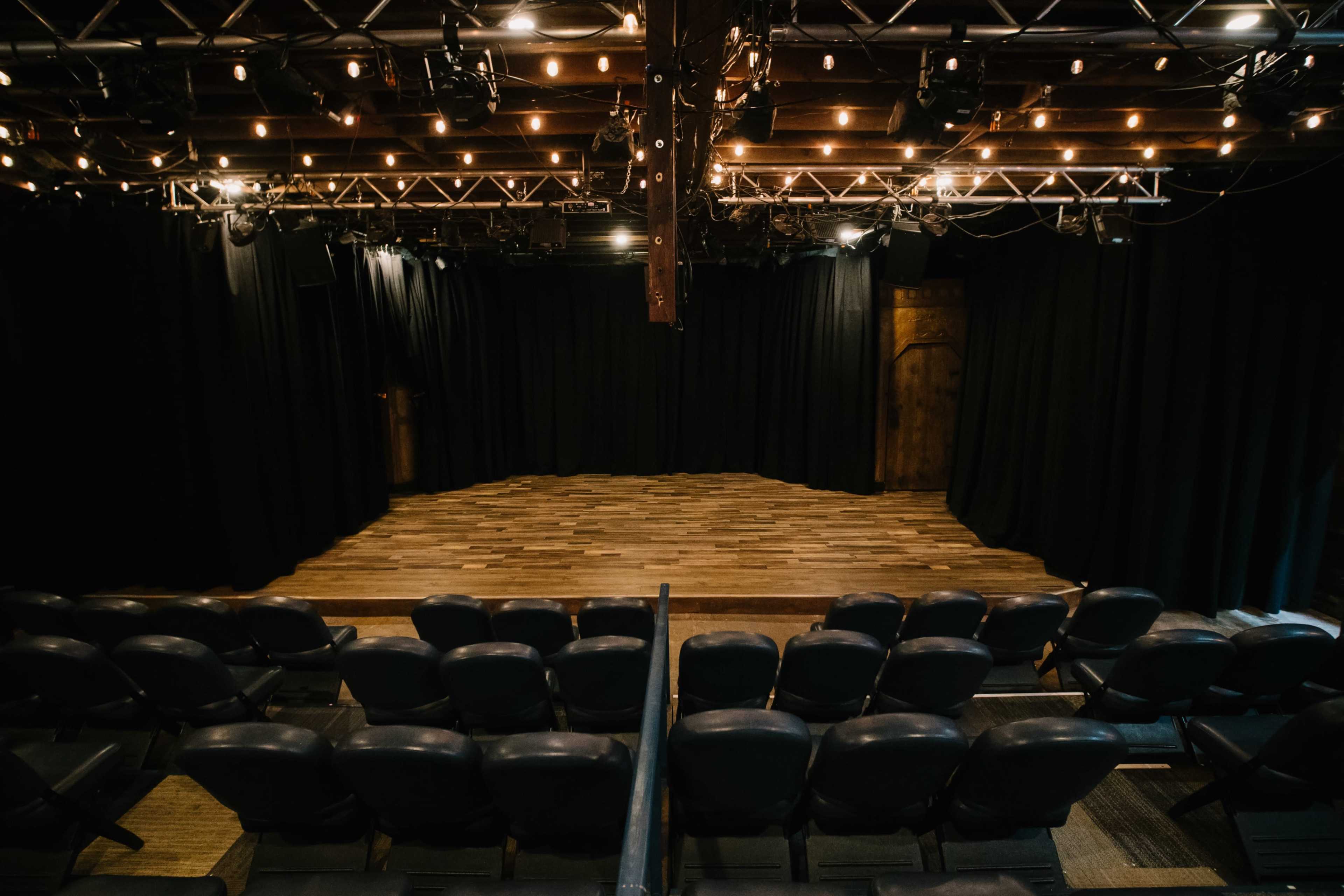 A theater stage is set with wooden flooring and surrounded by black curtains, with empty seats visible in the foreground.