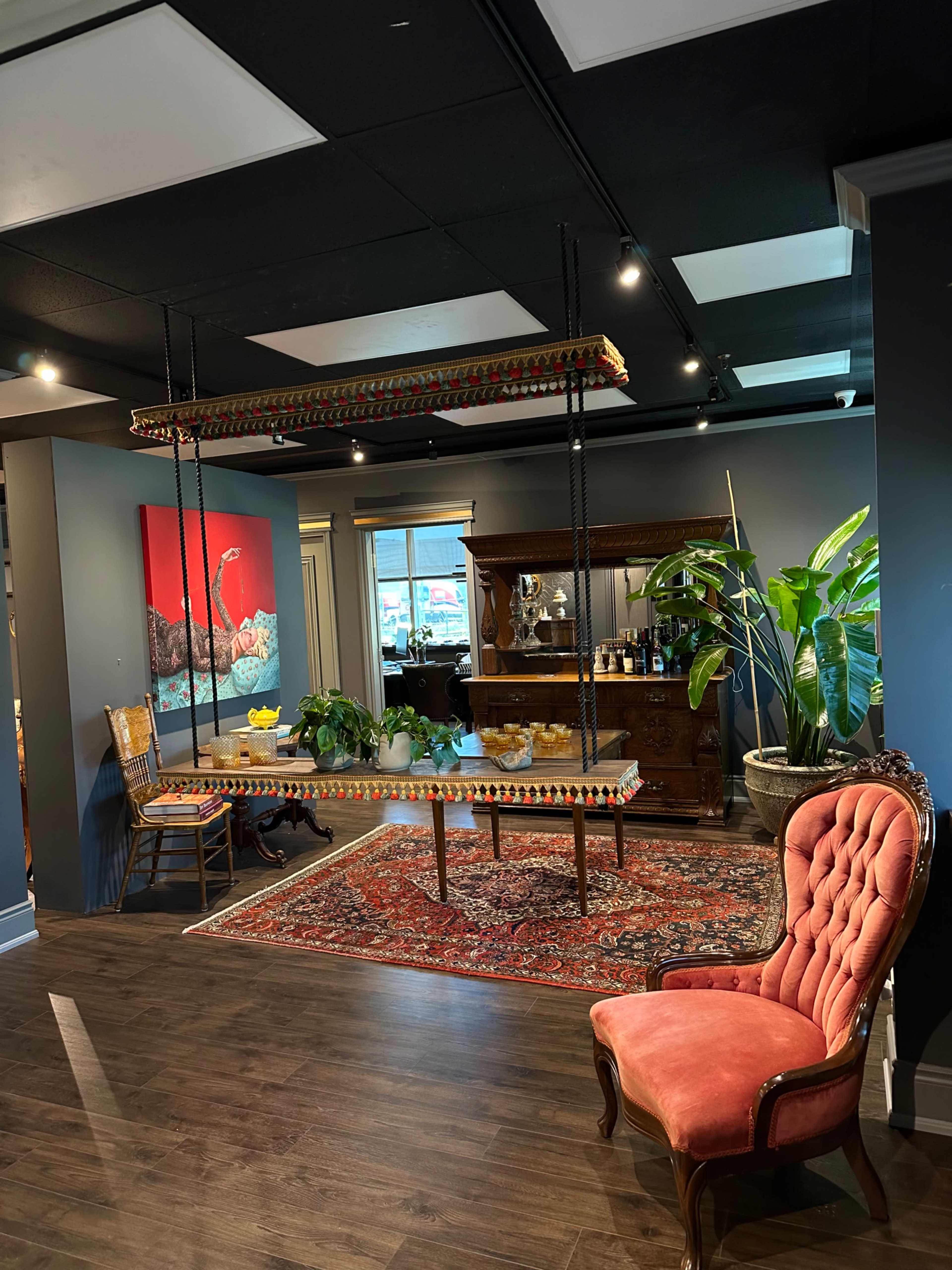 An inviting interior space featuring a hanging table with decorative edges, a vintage chair, a vibrant rug, and a bar area against a backdrop of dark blue walls and indoor plants.