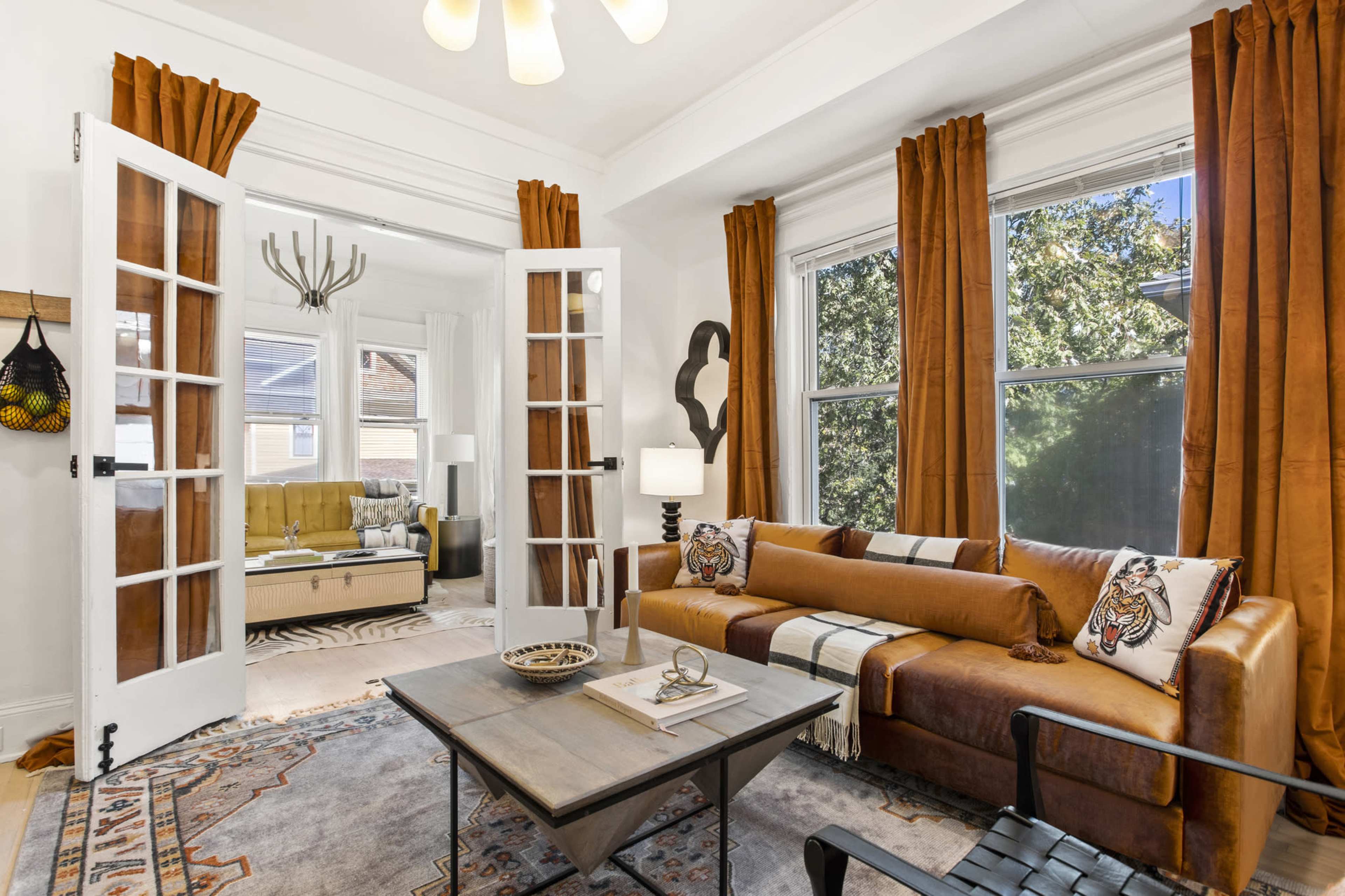 A cozy living room features a brown sofa, a coffee table, and large windows dressed with orange curtains, with a glimpse of a second room visible through French doors.