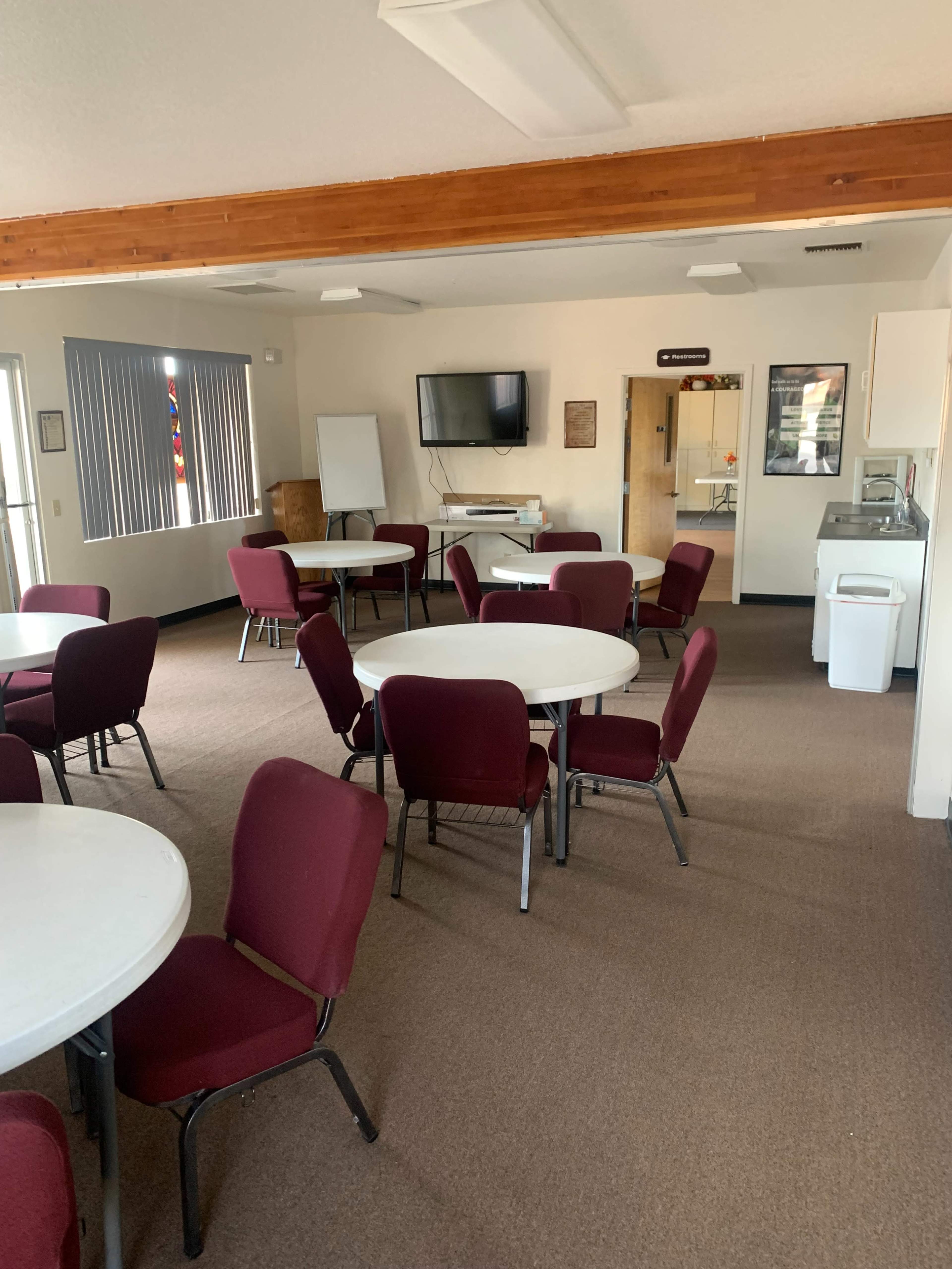 An empty room with several round tables and chairs arranged neatly, and a kitchenette area in the corner.
