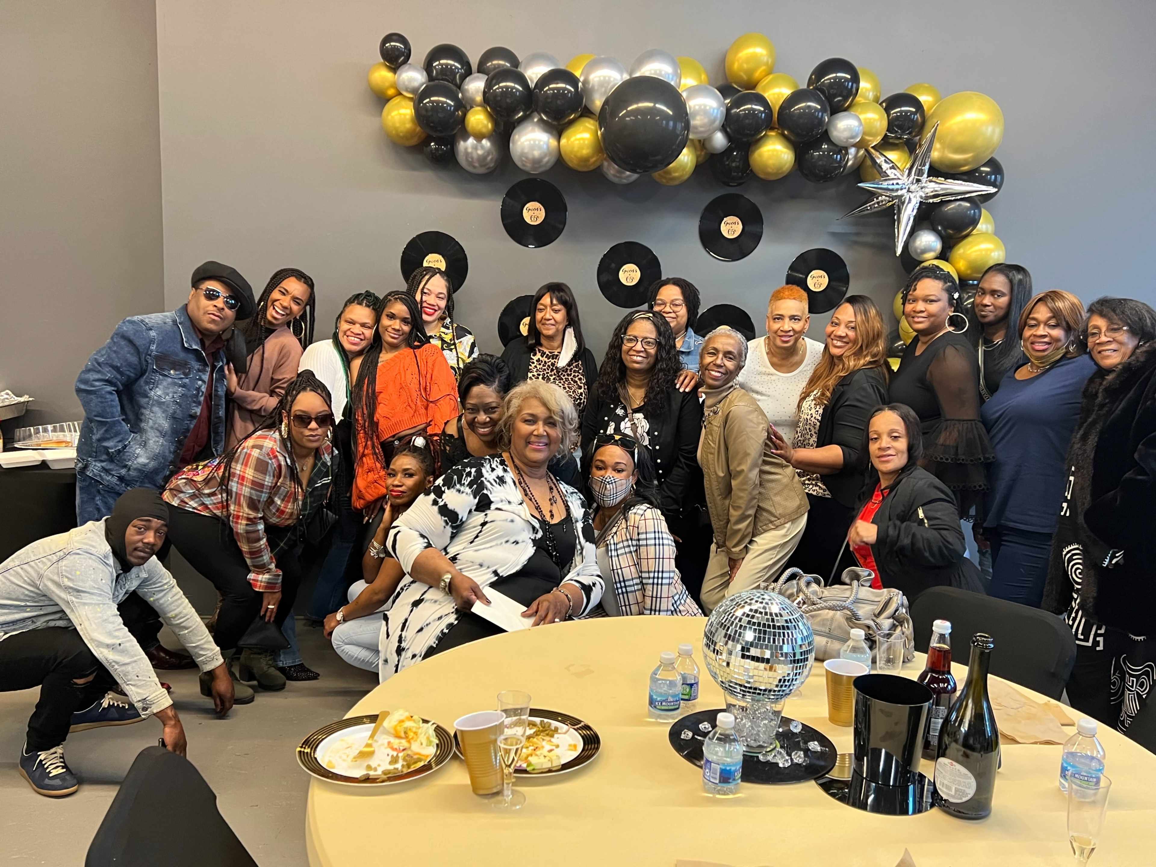 A diverse group of people gathers in a decorated room with a wall of balloons, records, and a disco ball, posing for a photo around a table with food and drinks.