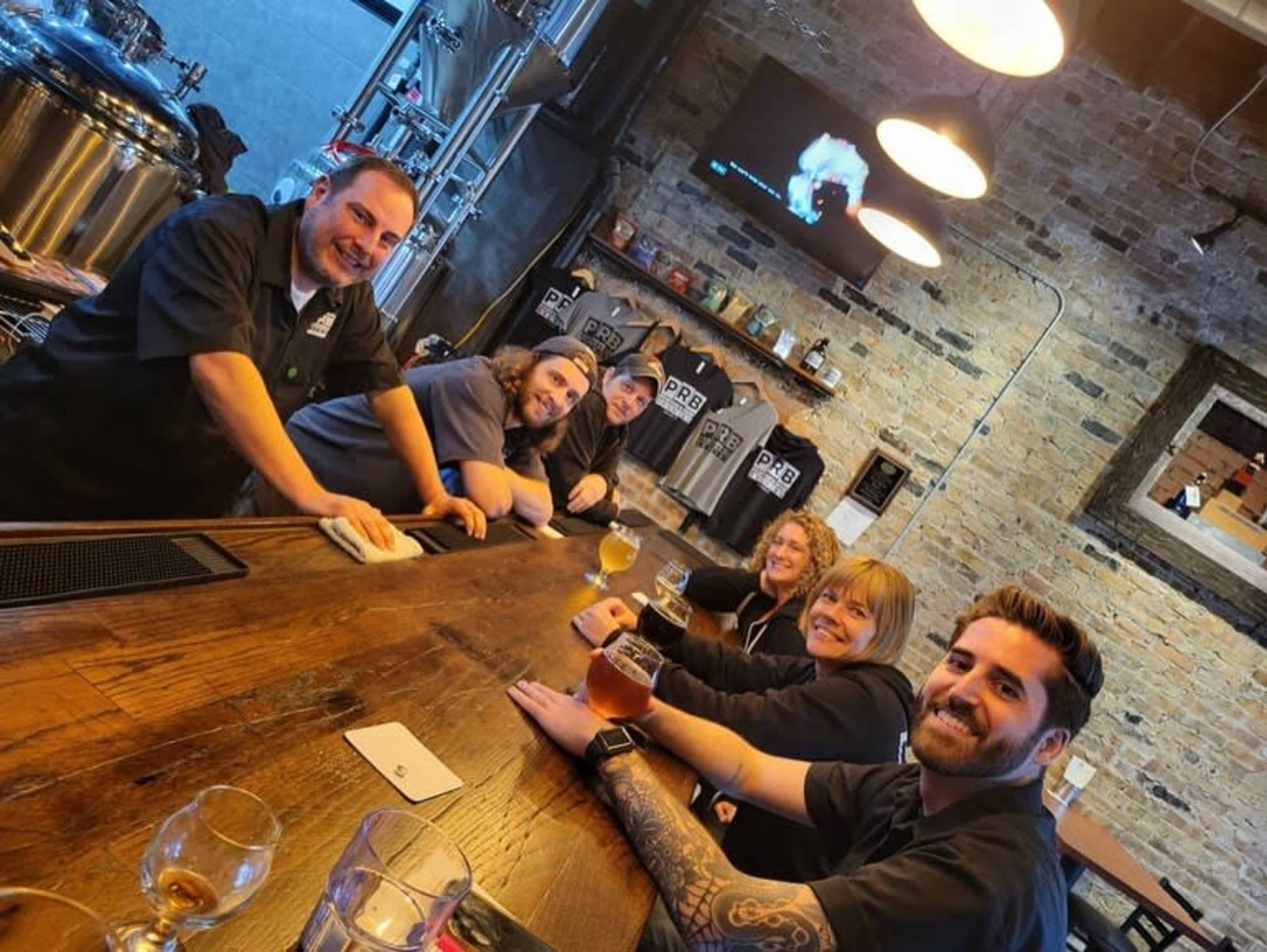 A group of six people sits at a wooden bar in a brewery, with brewing equipment visible in the background.