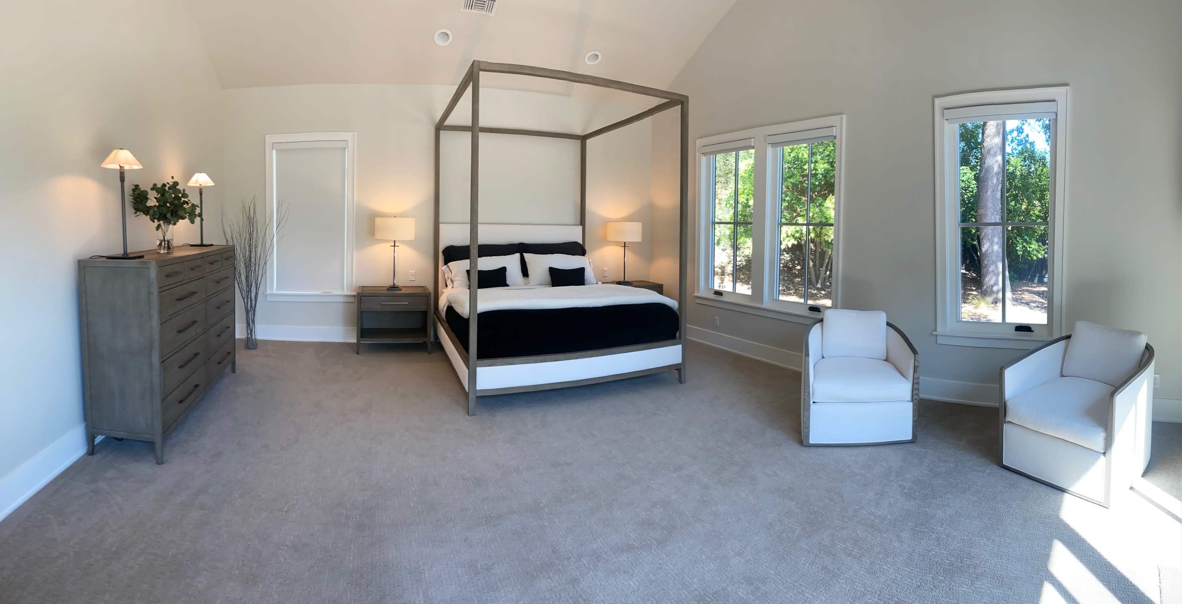 A spacious bedroom featuring a four-poster bed, a dresser, two modern armchairs, and large windows that let in natural light.