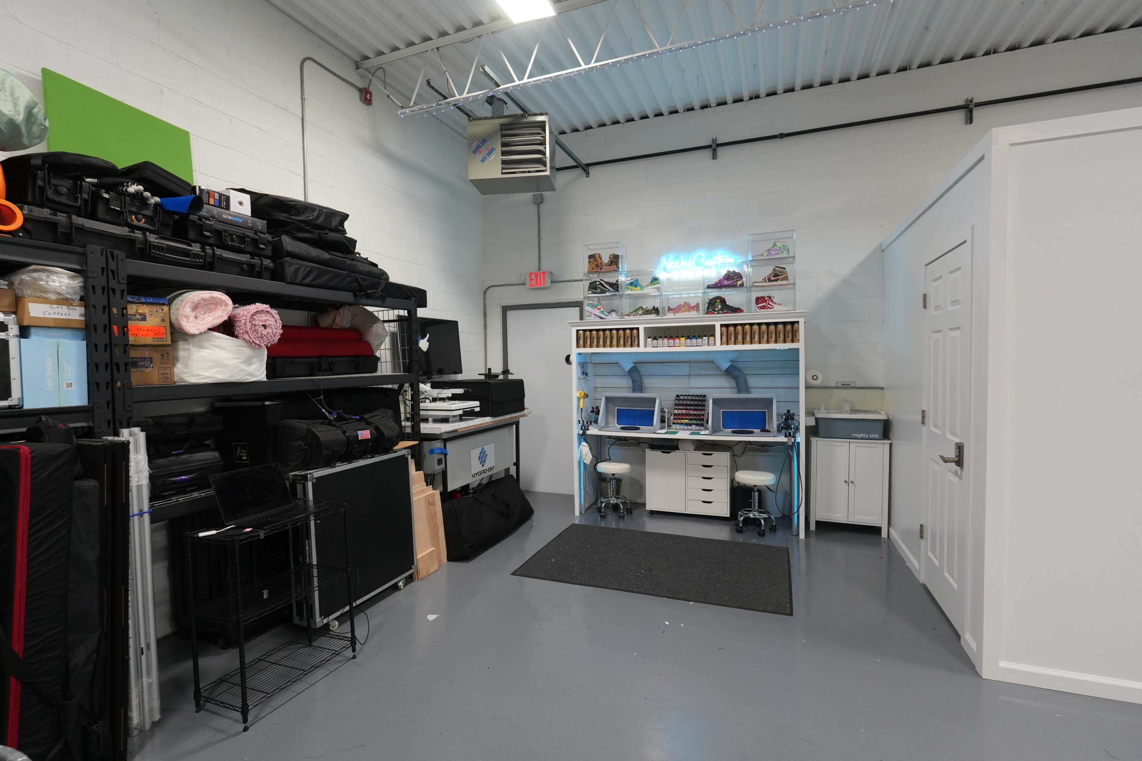 The image shows a spacious workspace with shelving units on the left filled with various equipment, while a designated desk area with blue accents and a neon sign is situated on the right alongside a closed door.