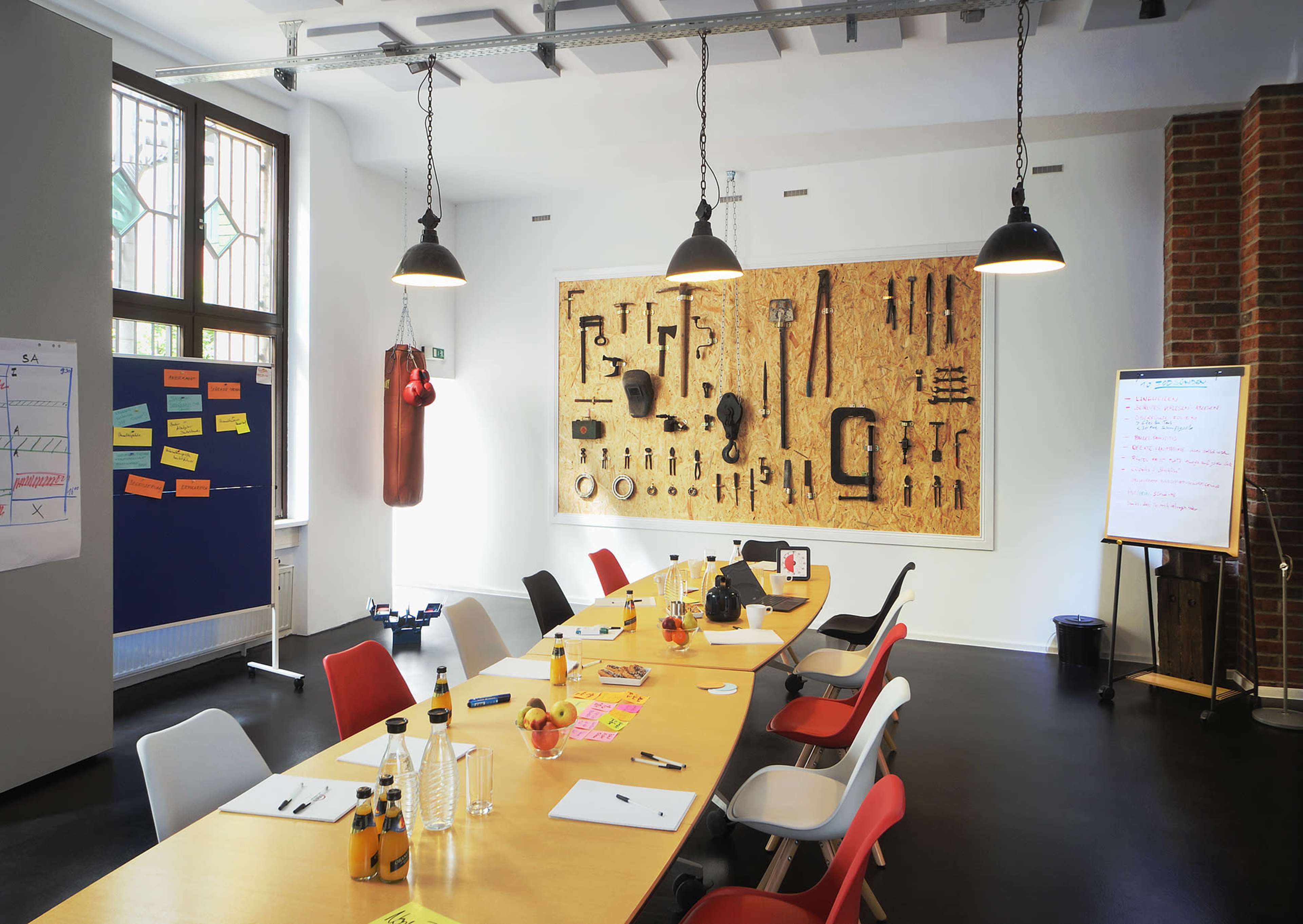 The image shows a modern meeting room with a long table set up for a workshop, featuring a wall display of various tools and a punching bag.