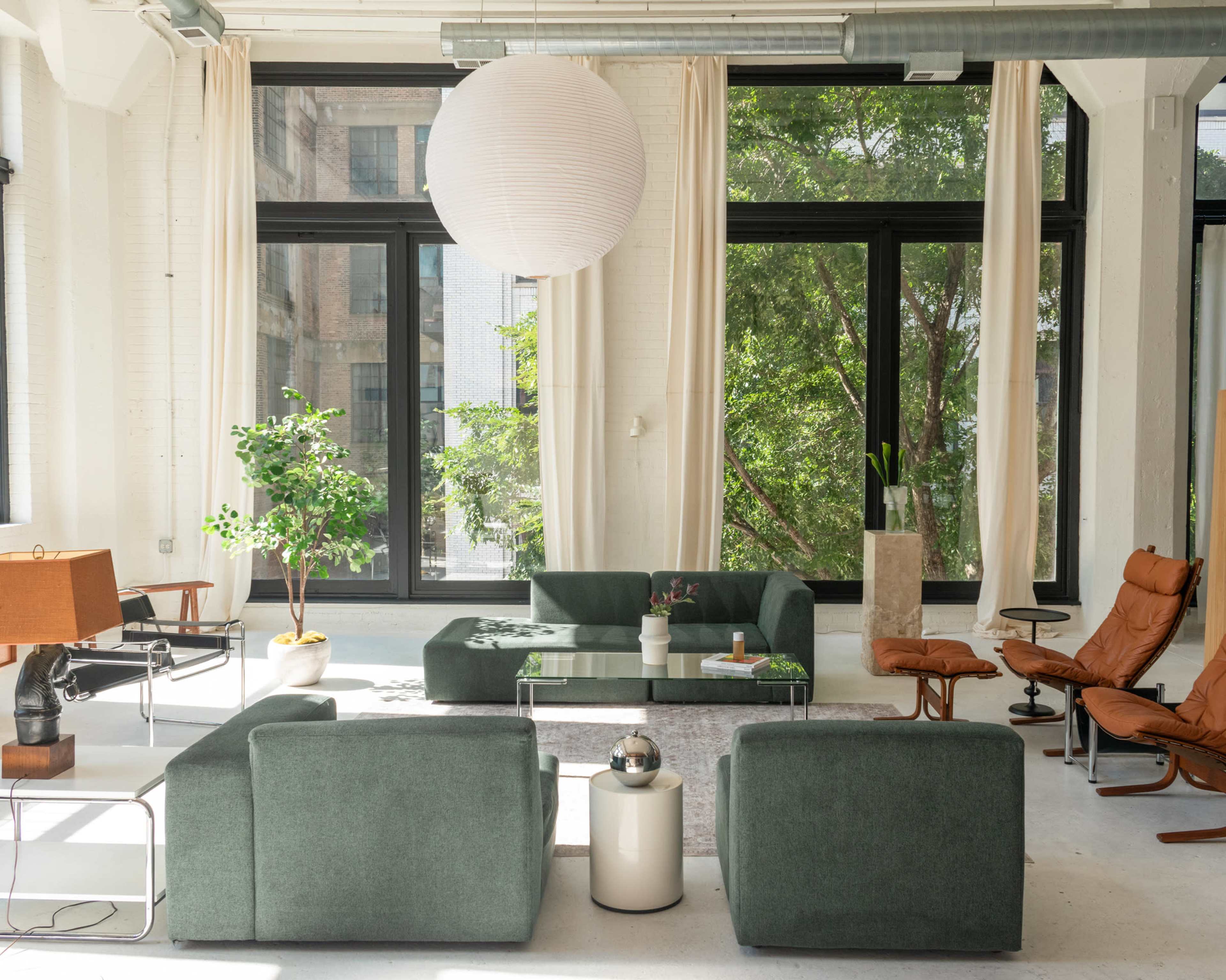 The room features a modern living space with large windows, green sofas, a glass coffee table, and a mix of wood and leather furniture.