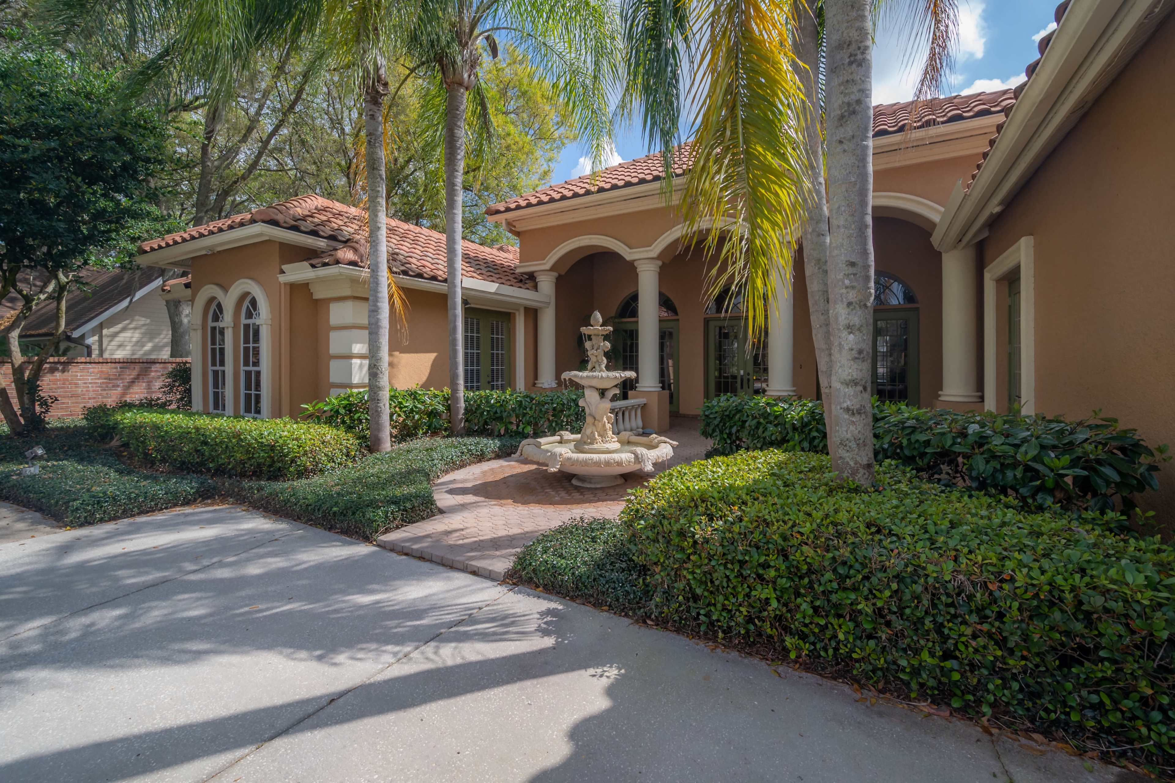 A residential home with a fountain in the front yard, surrounded by neatly trimmed hedges and palm trees.