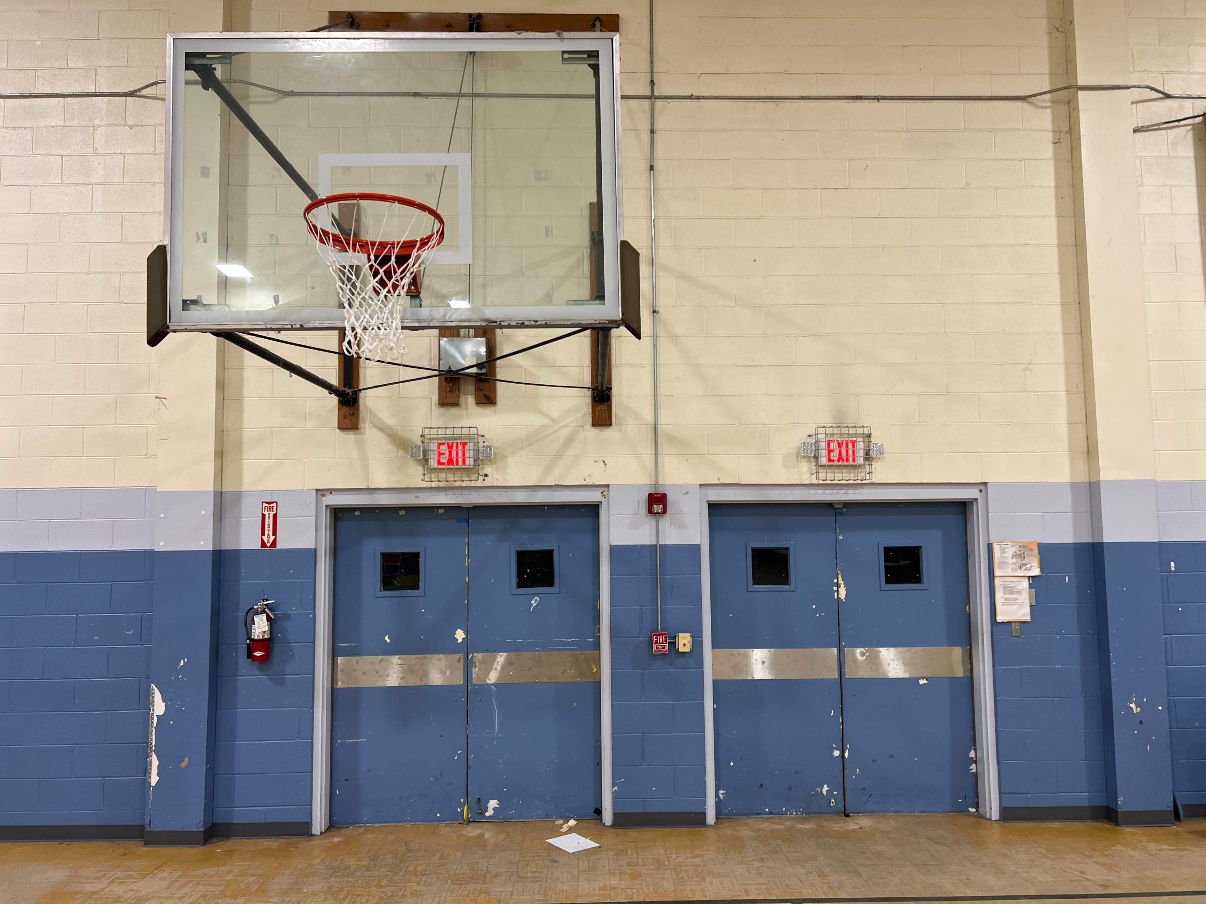 The image shows a basketball hoop mounted on a wall above two double doors in a gymnasium, with worn blue and white paint on the walls.