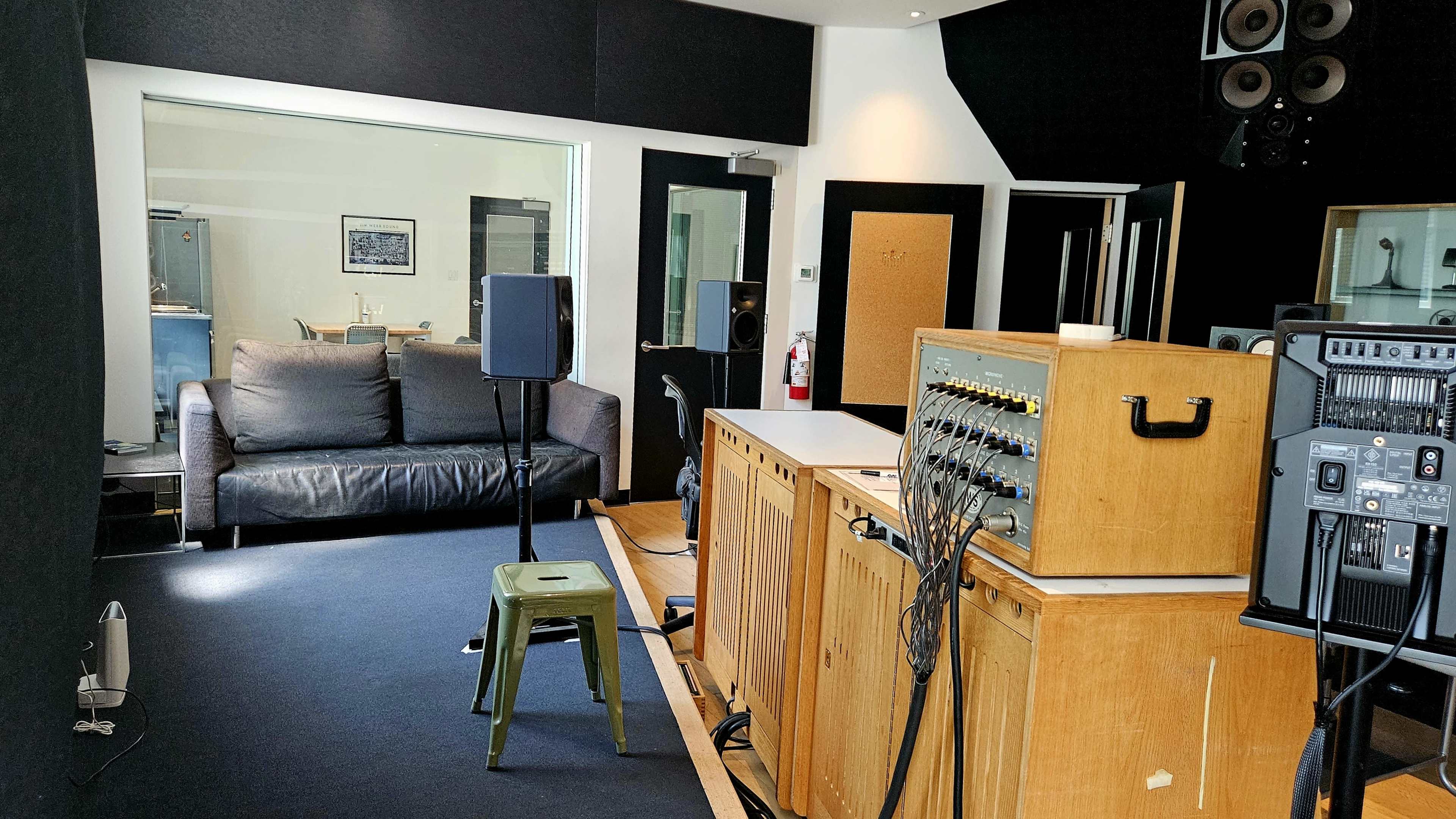 The image shows a recording studio with a couch, a sound mixing console, speakers, and a view into another room through a glass wall.