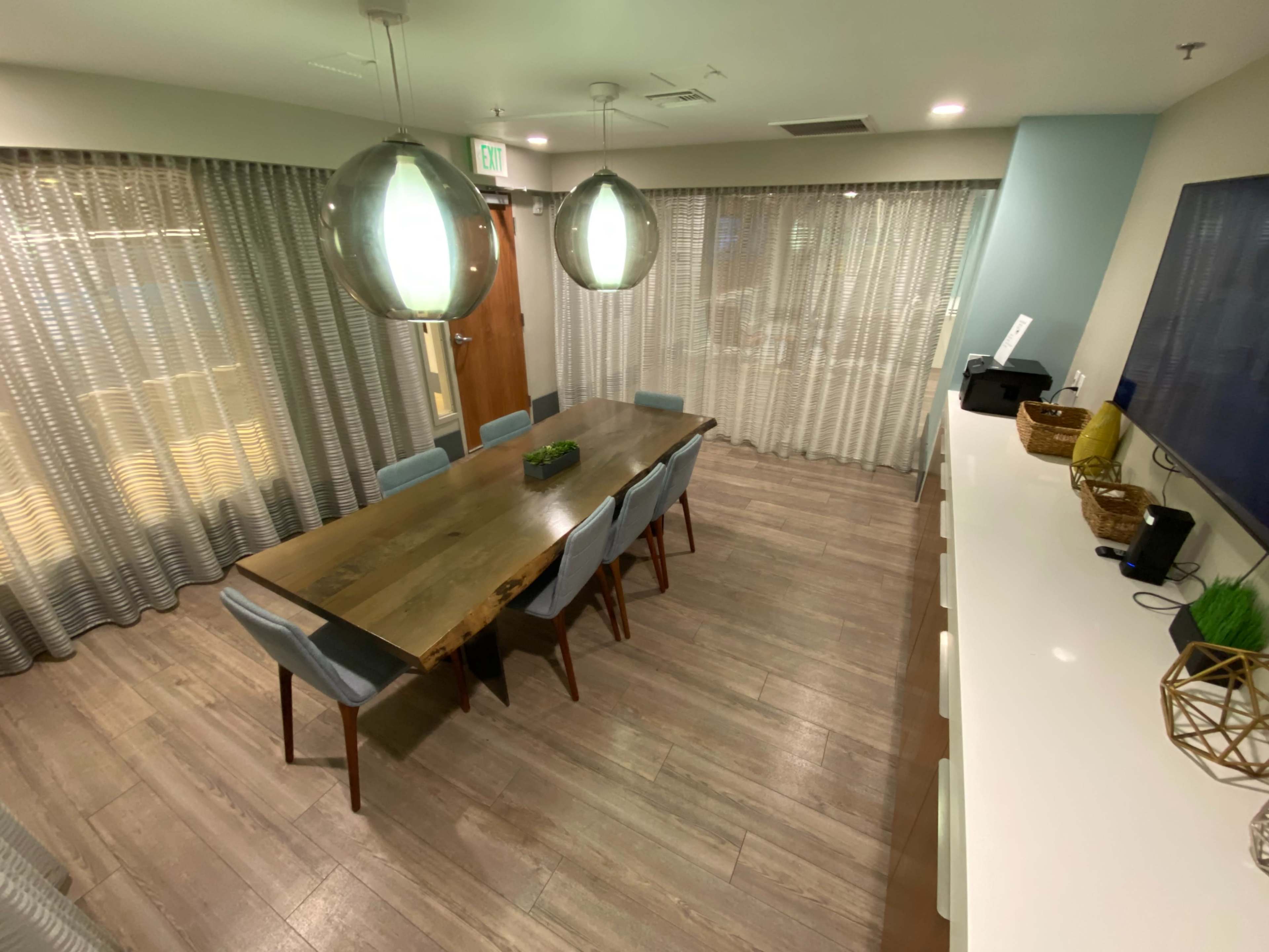 A modern conference room featuring a large wooden table surrounded by chairs, with pendant lights above and sheer curtains covering the windows.