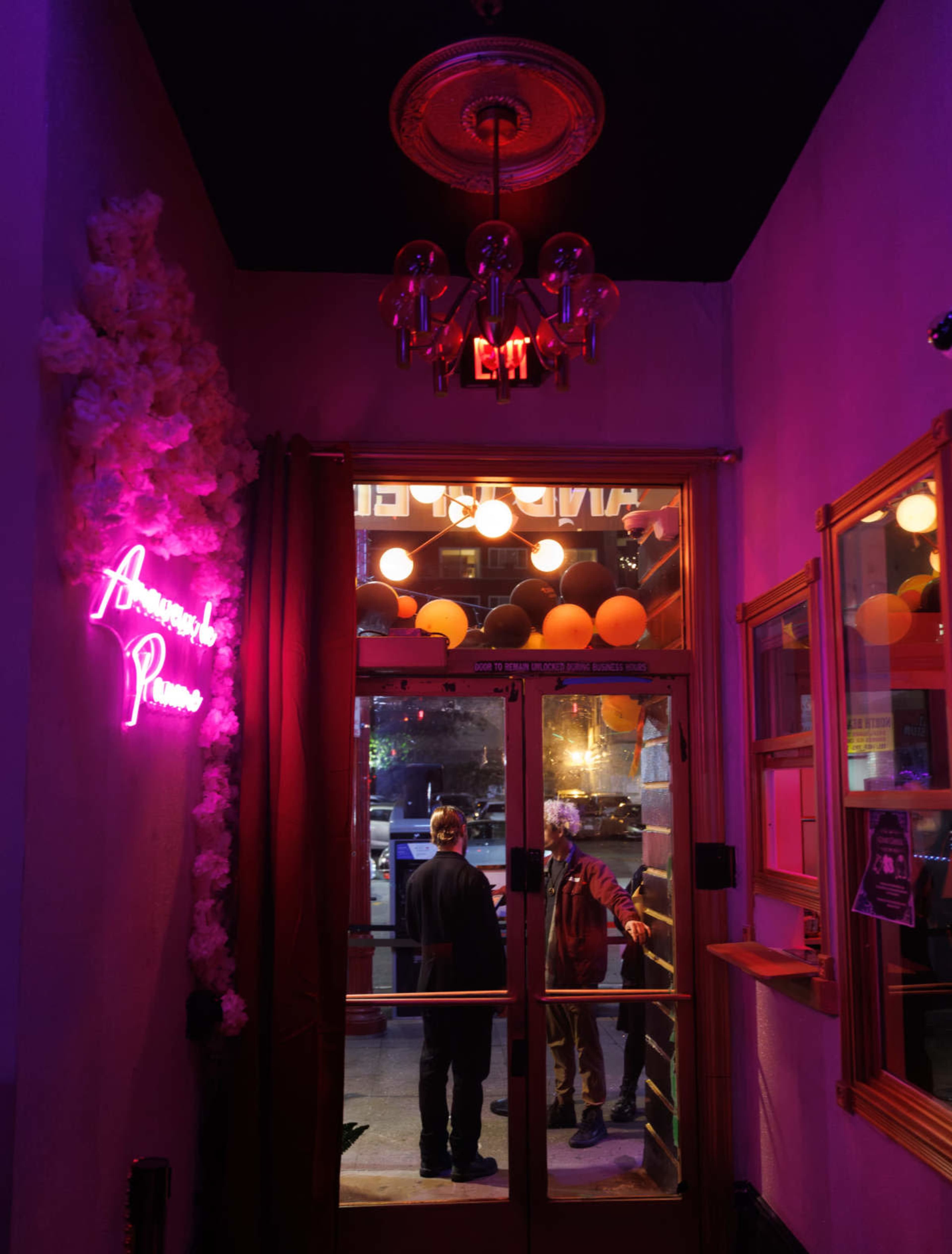 The image shows an entrance to a venue with a neon sign and a chandelier, illuminated by soft pink and orange lighting.
