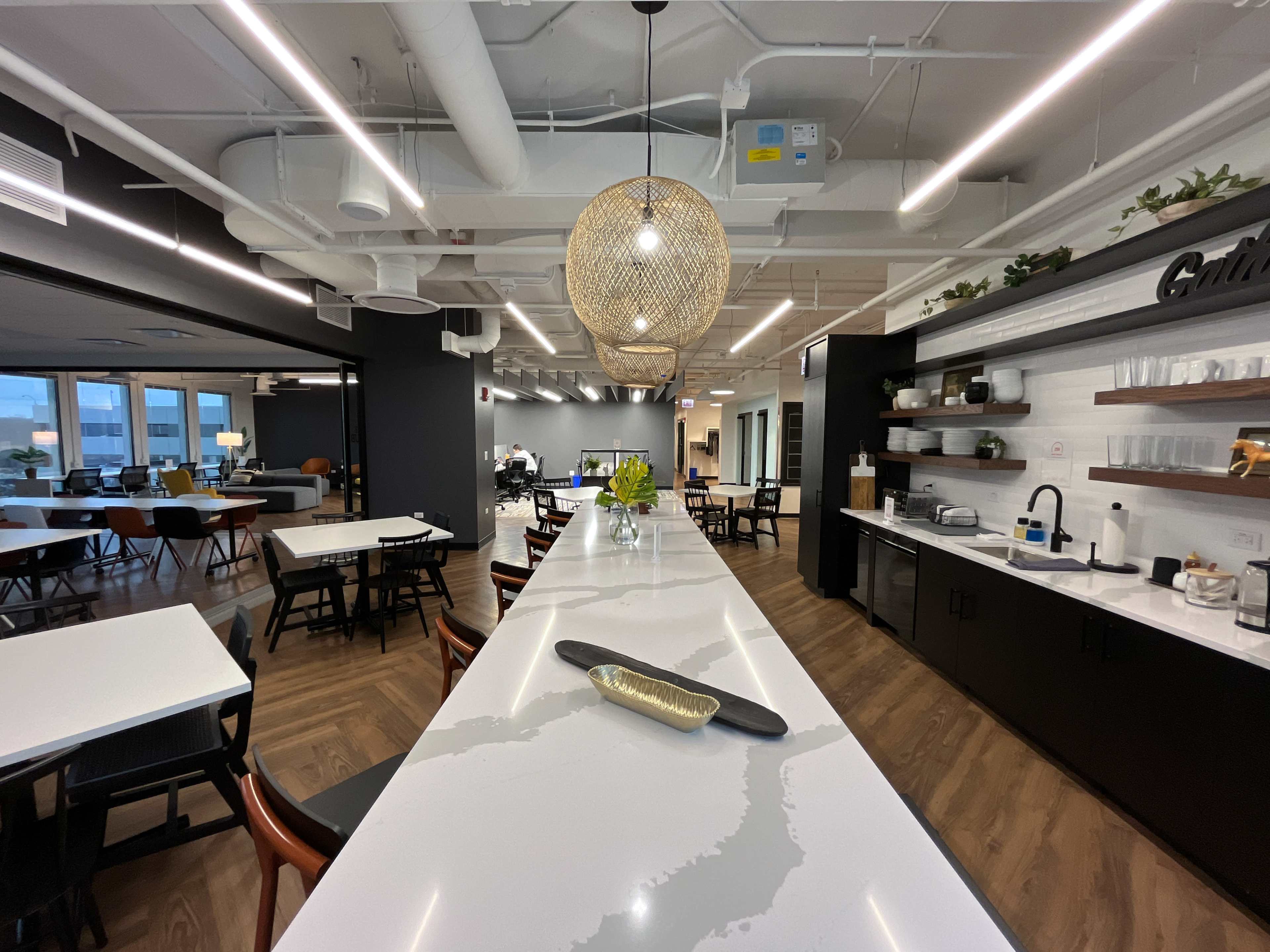 The image shows a modern office kitchen with a long white countertop, decorative pendant lights, and various seating areas in the background.