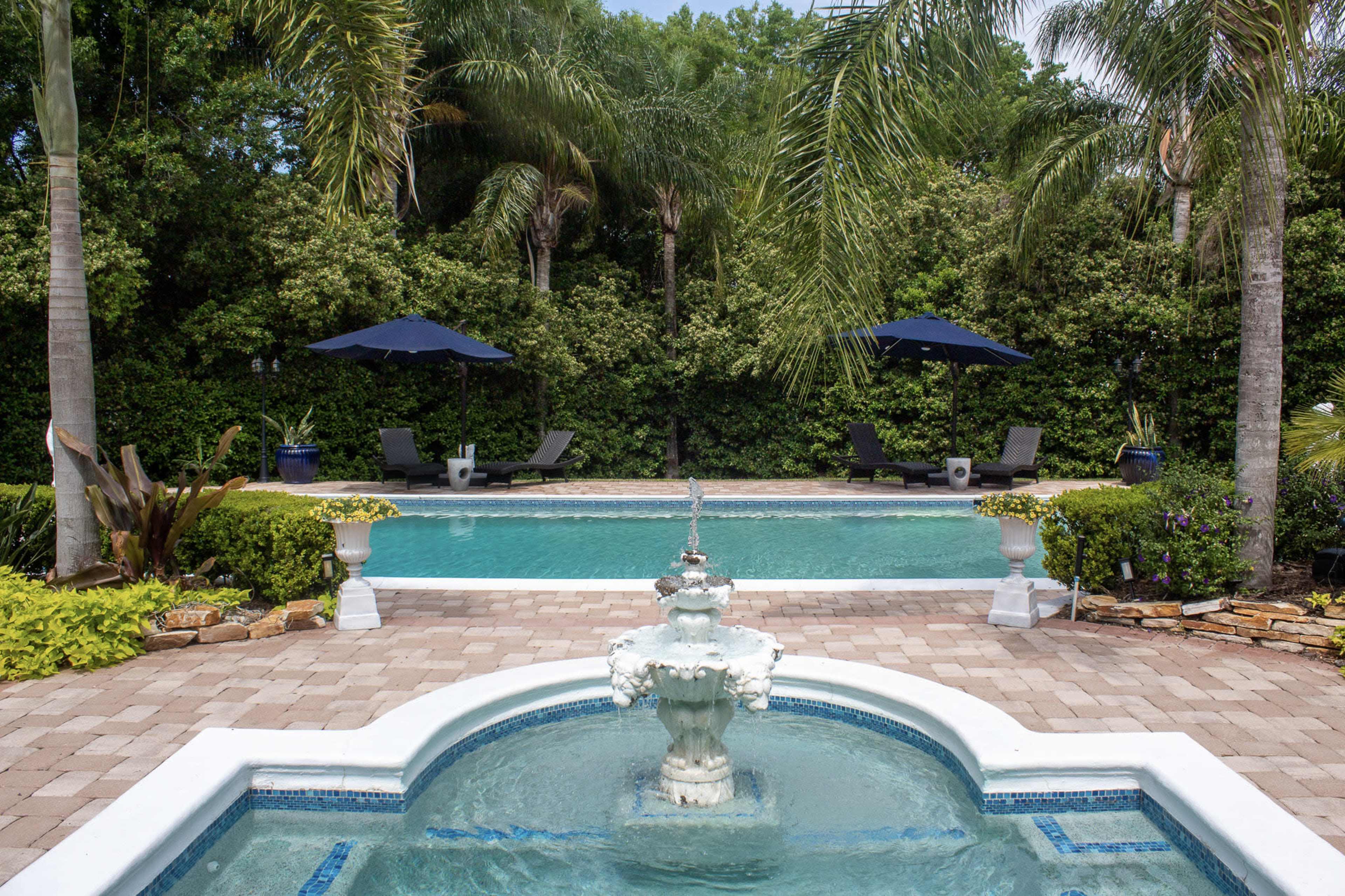 A swimming pool surrounded by palm trees, lounge chairs, and two blue umbrellas, with a decorative fountain in the foreground.