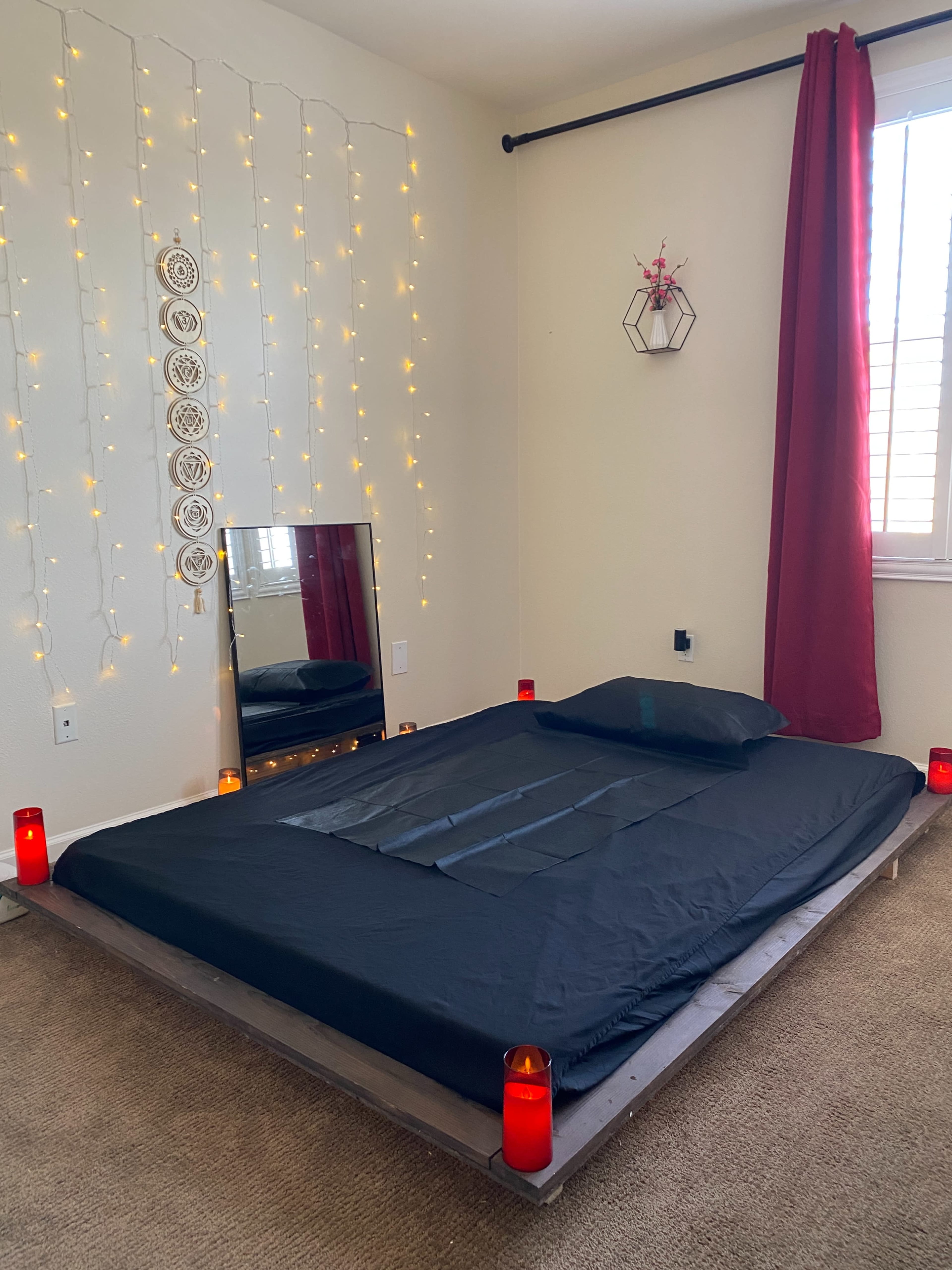 A minimalist bedroom features a low black bed on a wooden platform, surrounded by lit candles and fairy lights, with a large mirror and a red curtain by the window.