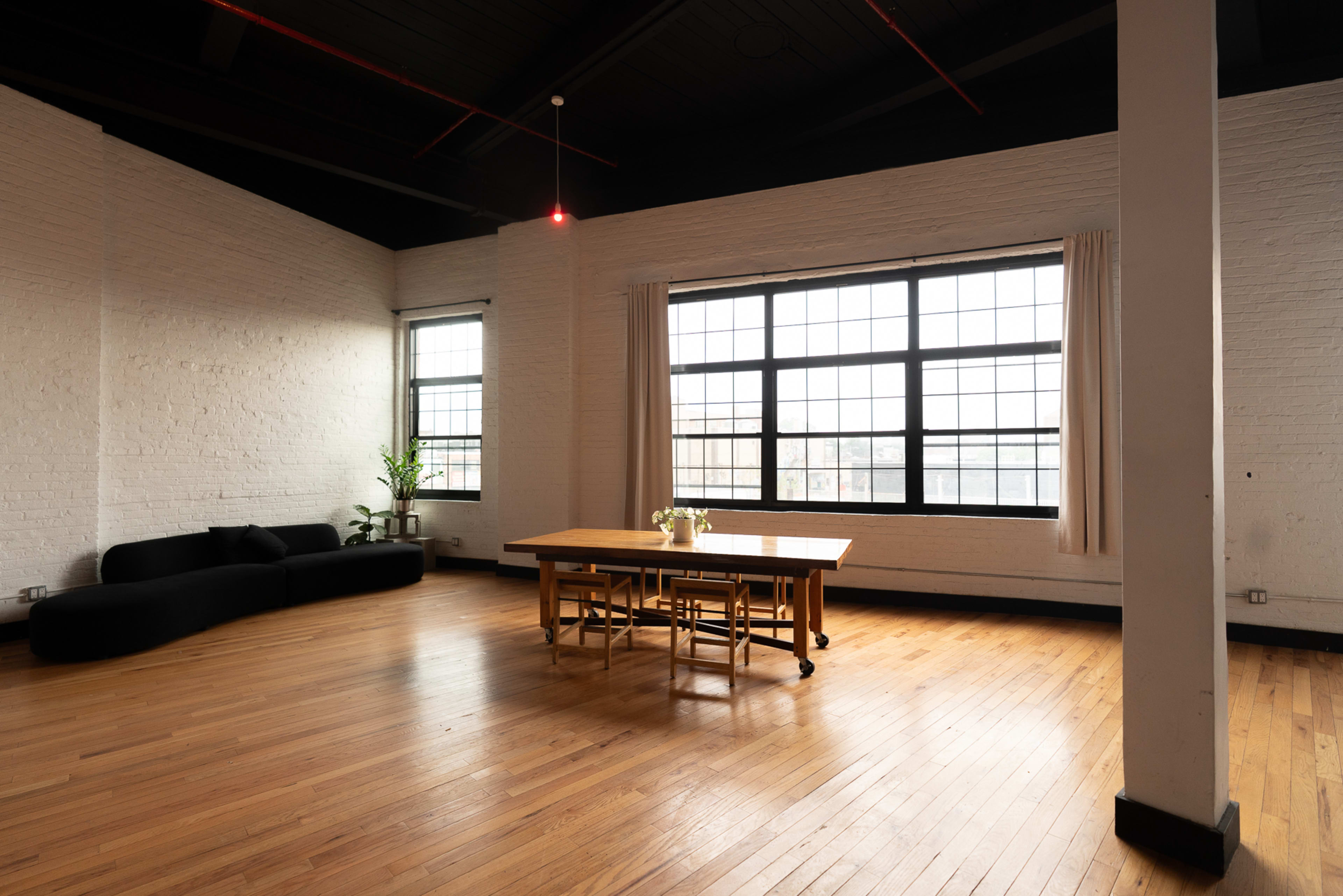 The image shows a spacious room with wooden flooring, a black couch, a wooden dining table, and large windows allowing natural light to enter.