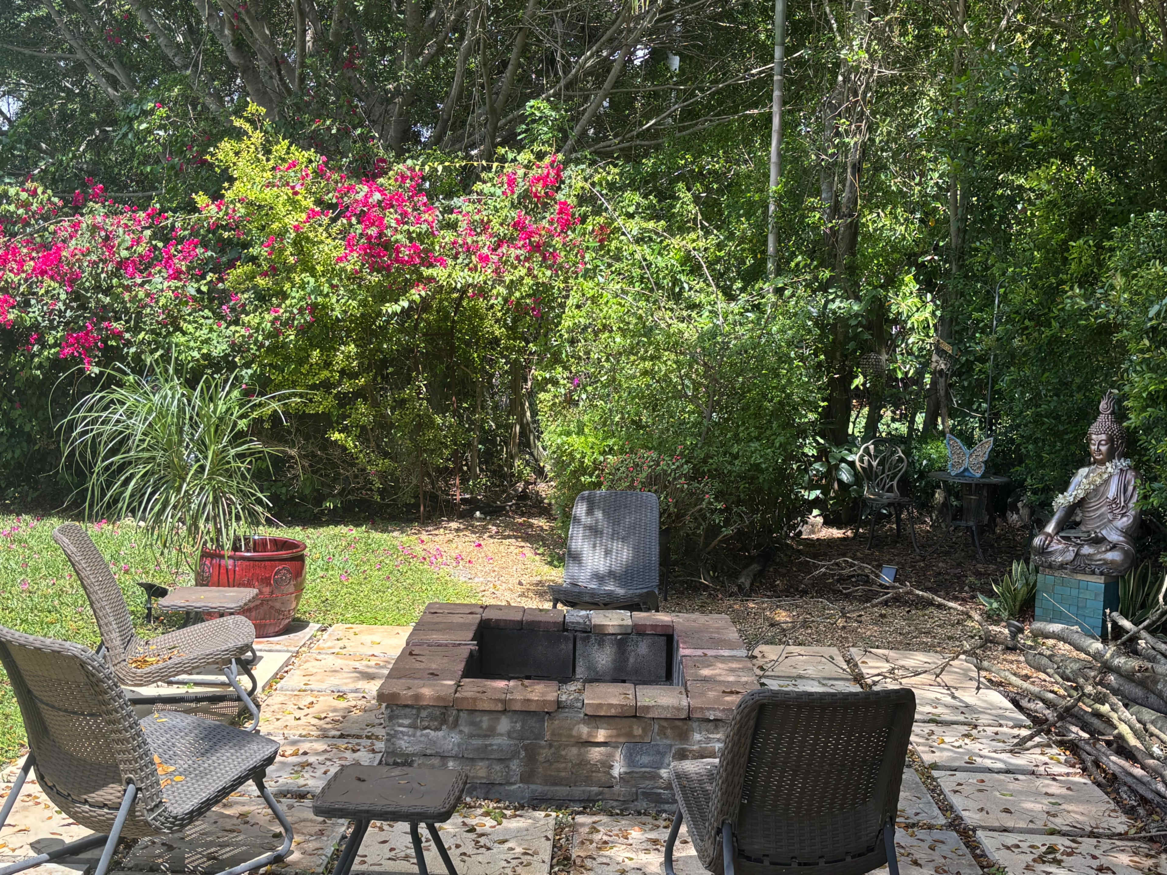 The image shows a backyard patio with a stone fire pit surrounded by several chairs, colorful plants, and a statue.