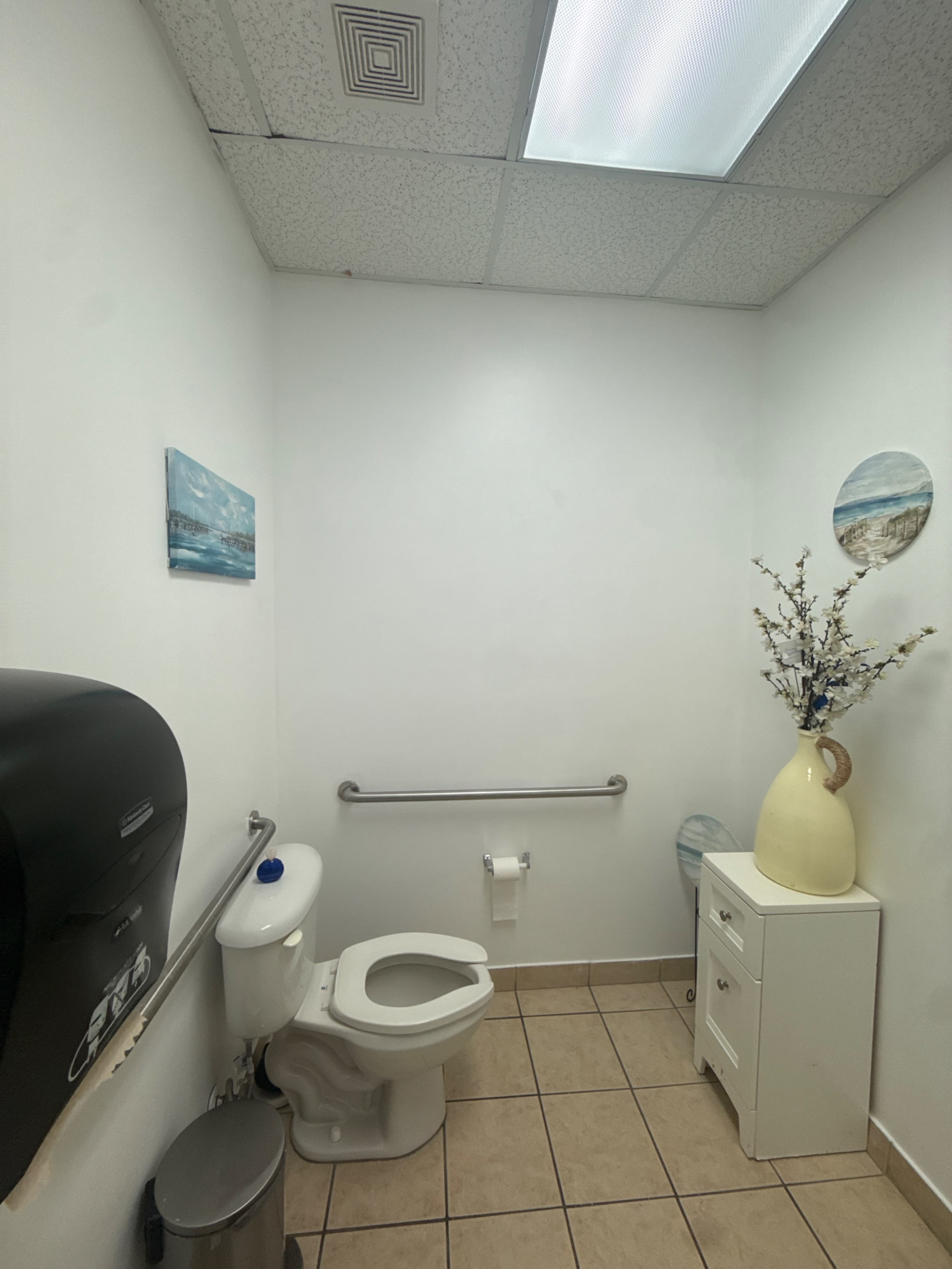 The image shows a clean restroom with a toilet, hand dryer, toilet paper holder, and decorative elements including a vase and wall art.