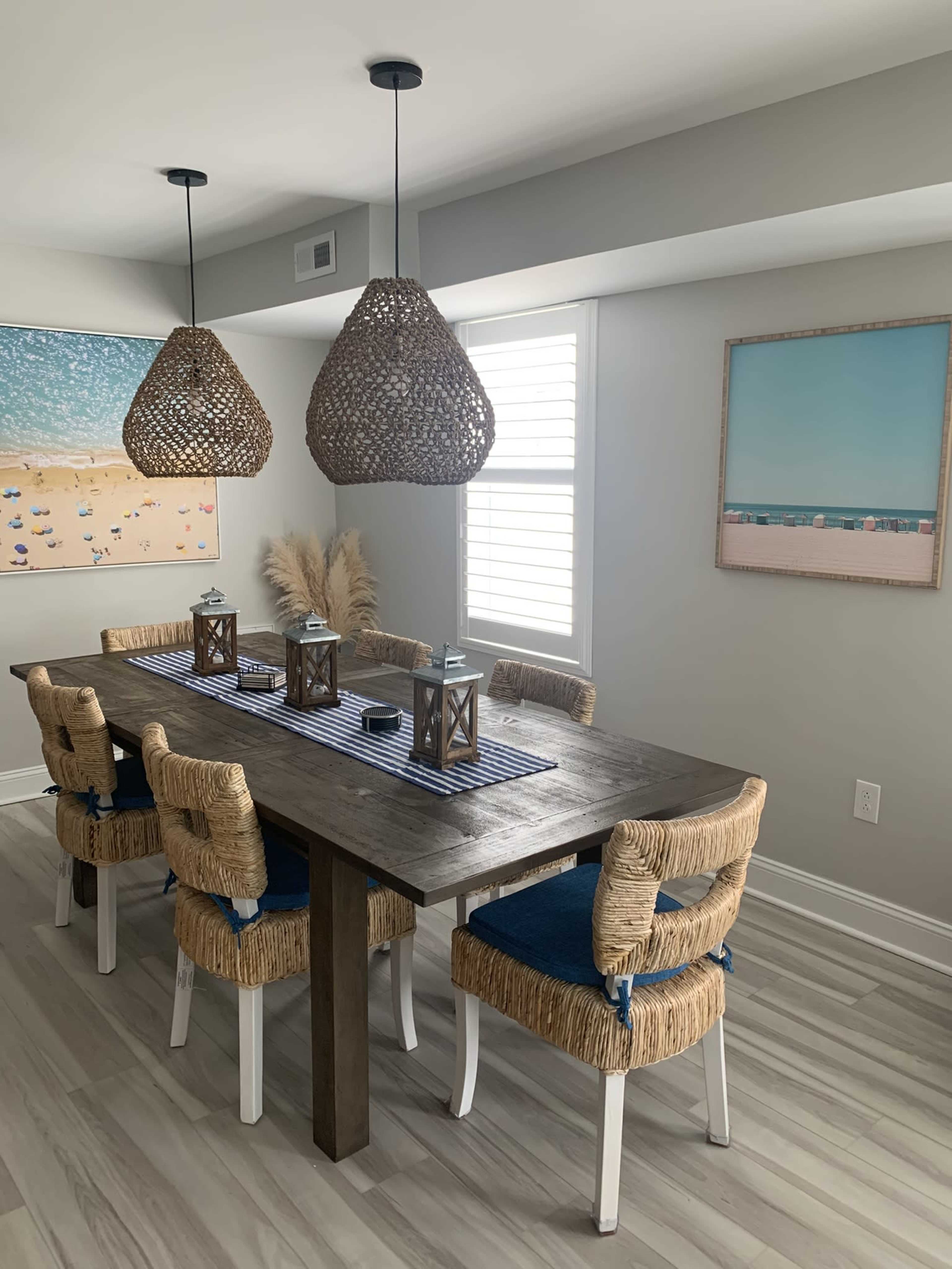 A modern dining room features a wooden table surrounded by woven chairs, two large pendant lights, and artwork on the walls.