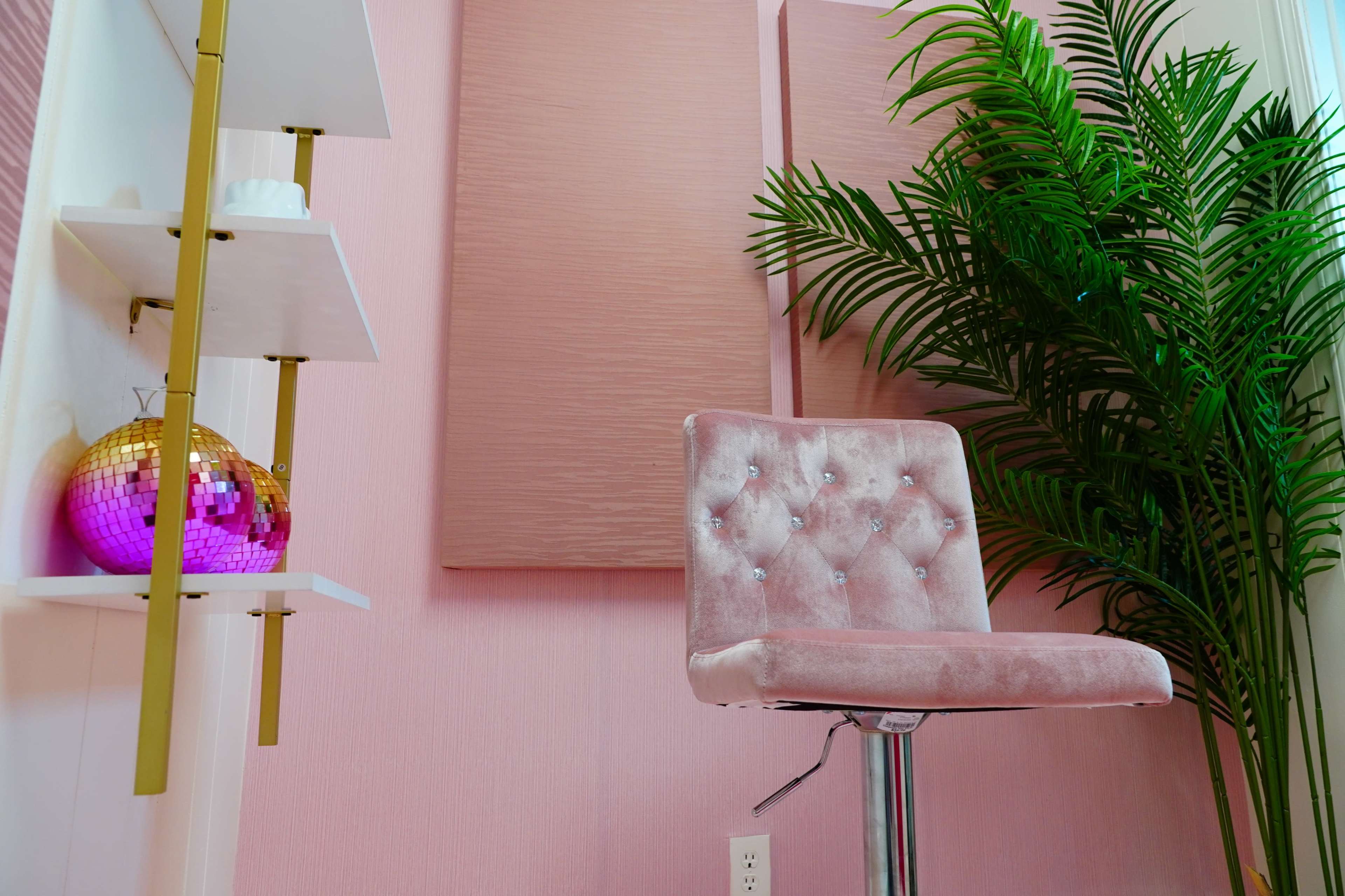 A pink velvet chair next to a gold and pink disco ball, accompanied by a tall green plant and pink wall panels.