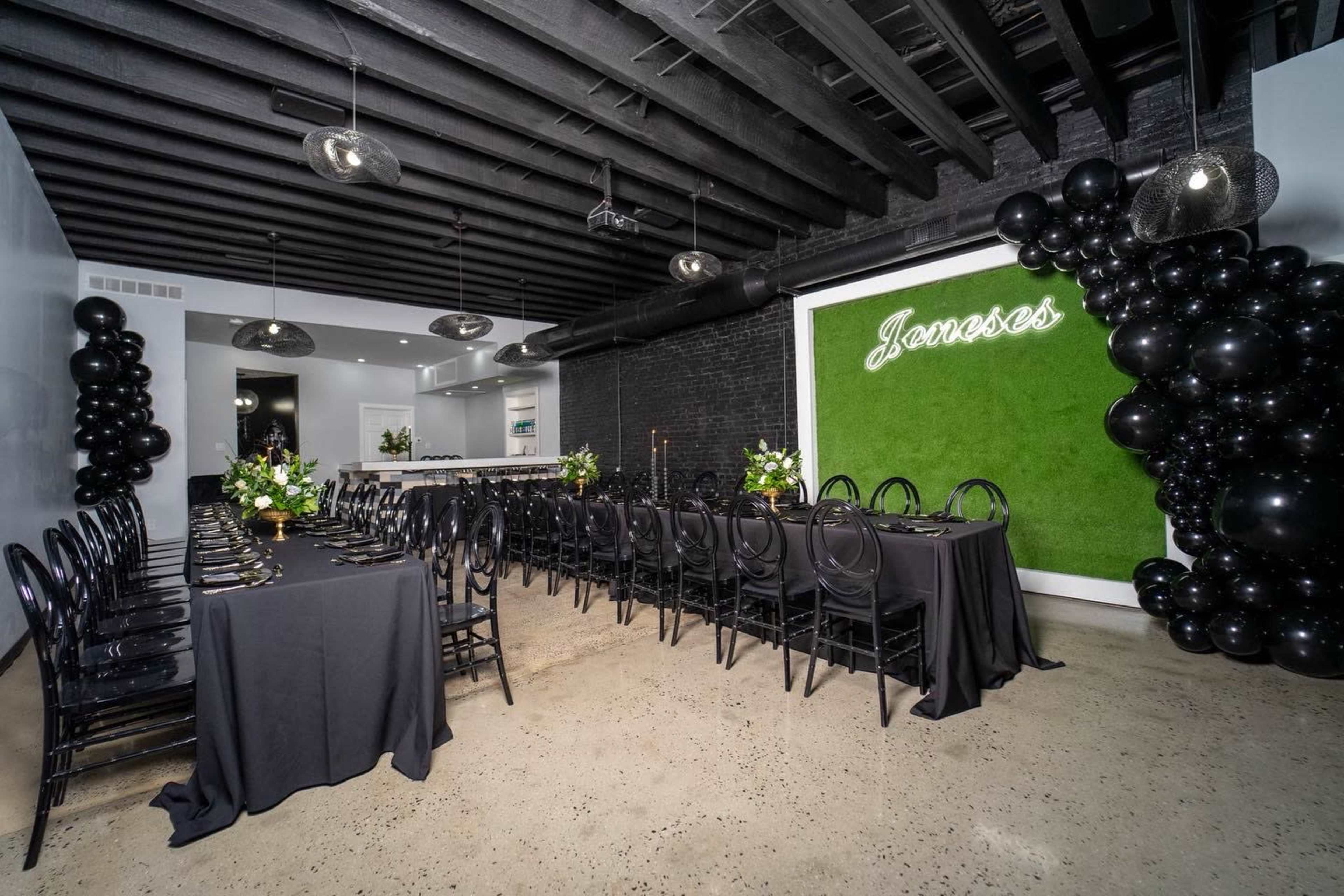 A spacious event room features a long black table set with dark chairs, adorned with floral centerpieces, and a green wall with illuminated signage, accented by black balloon decor.
