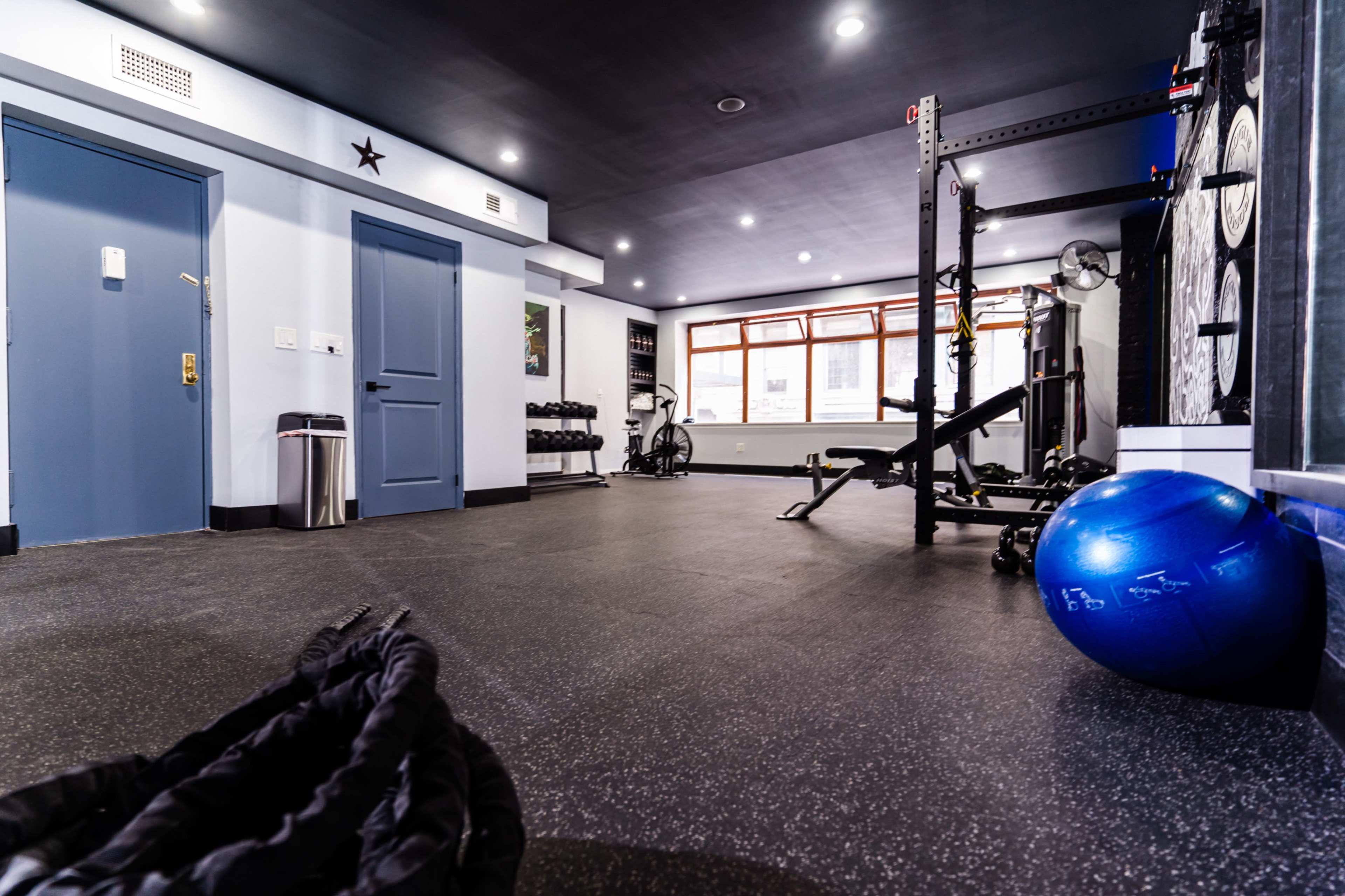 A fitness room with exercise equipment, including weights, a bench, and a stability ball, along with large windows providing natural light.