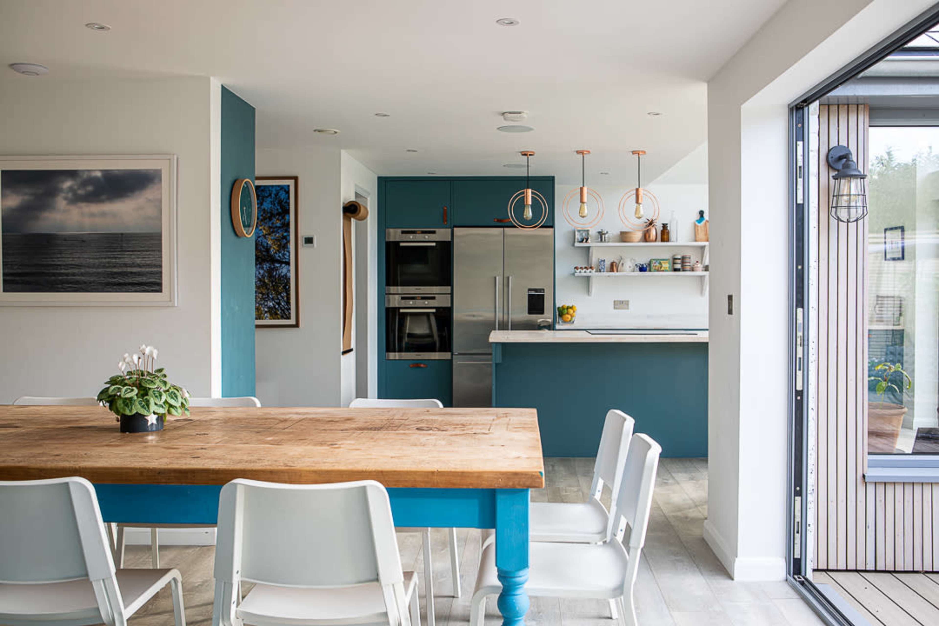 A modern kitchen and dining area featuring a wooden table surrounded by white chairs, with teal cabinetry and appliances in a well-lit space.