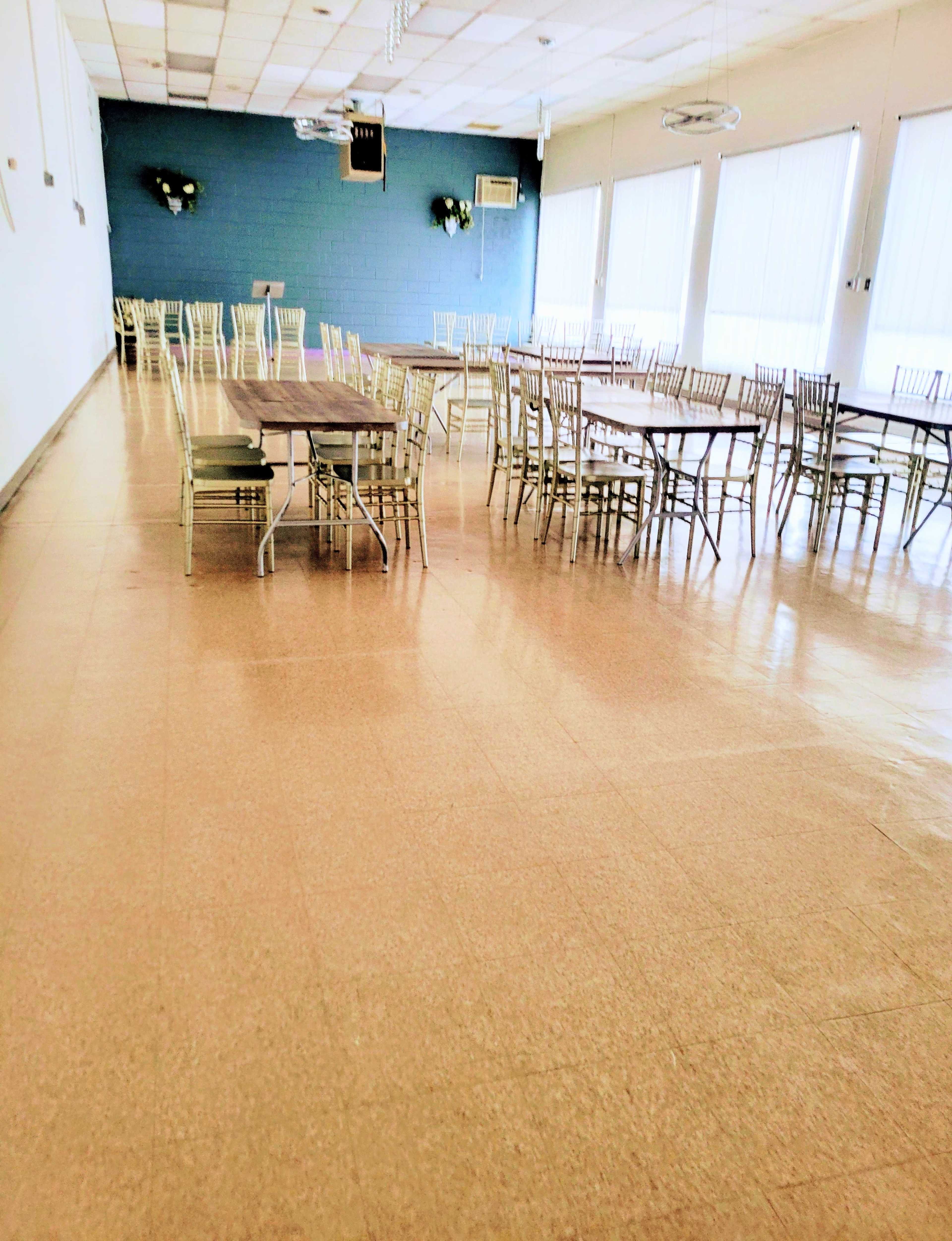 A spacious room features multiple rows of tables and chairs set against light-colored walls and large windows.