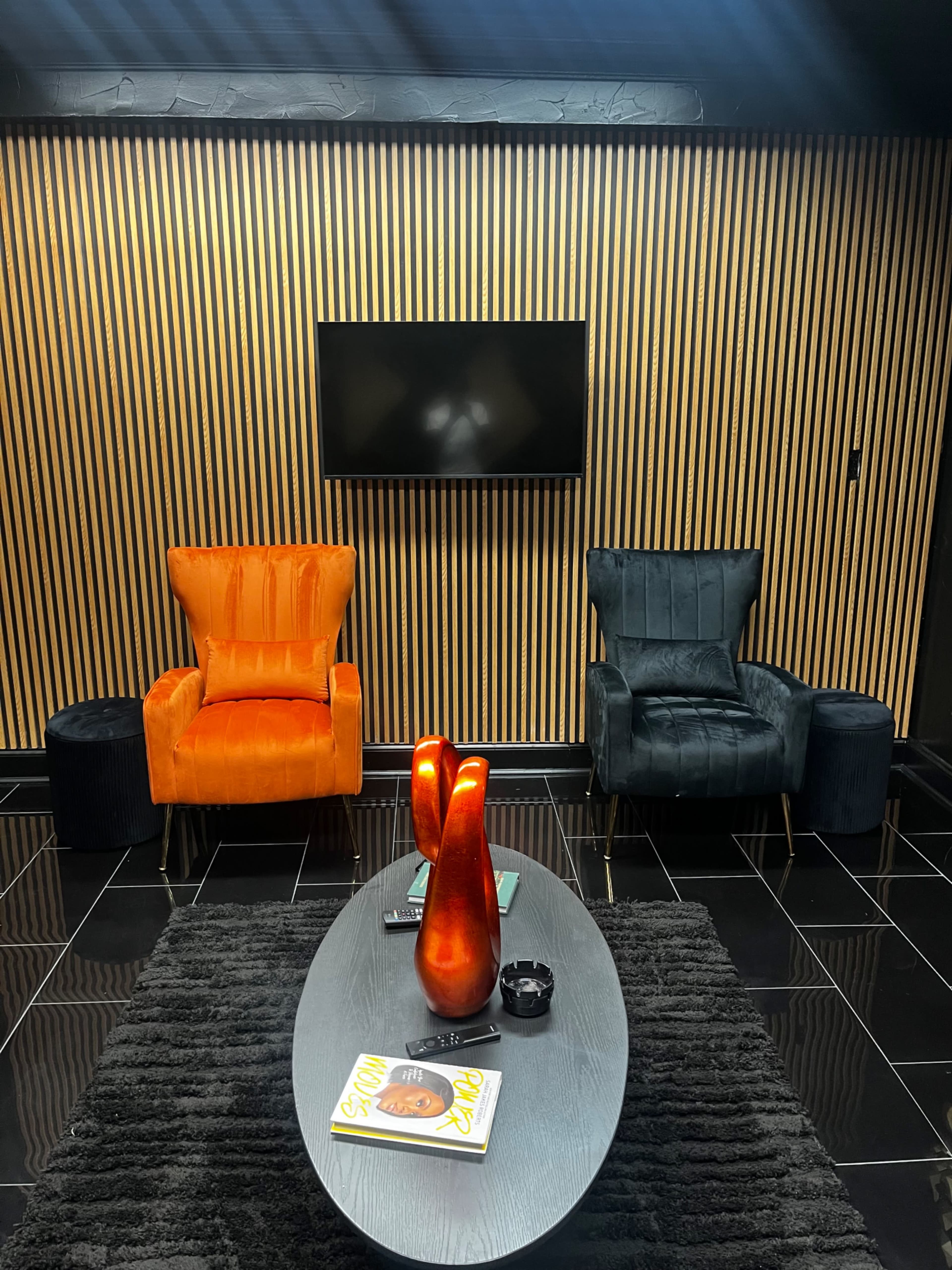 The image features two armchairs—one orange and one dark green—facing a flat-screen TV, with a coffee table and decorative items in a stylishly designed room with vertical striped walls.