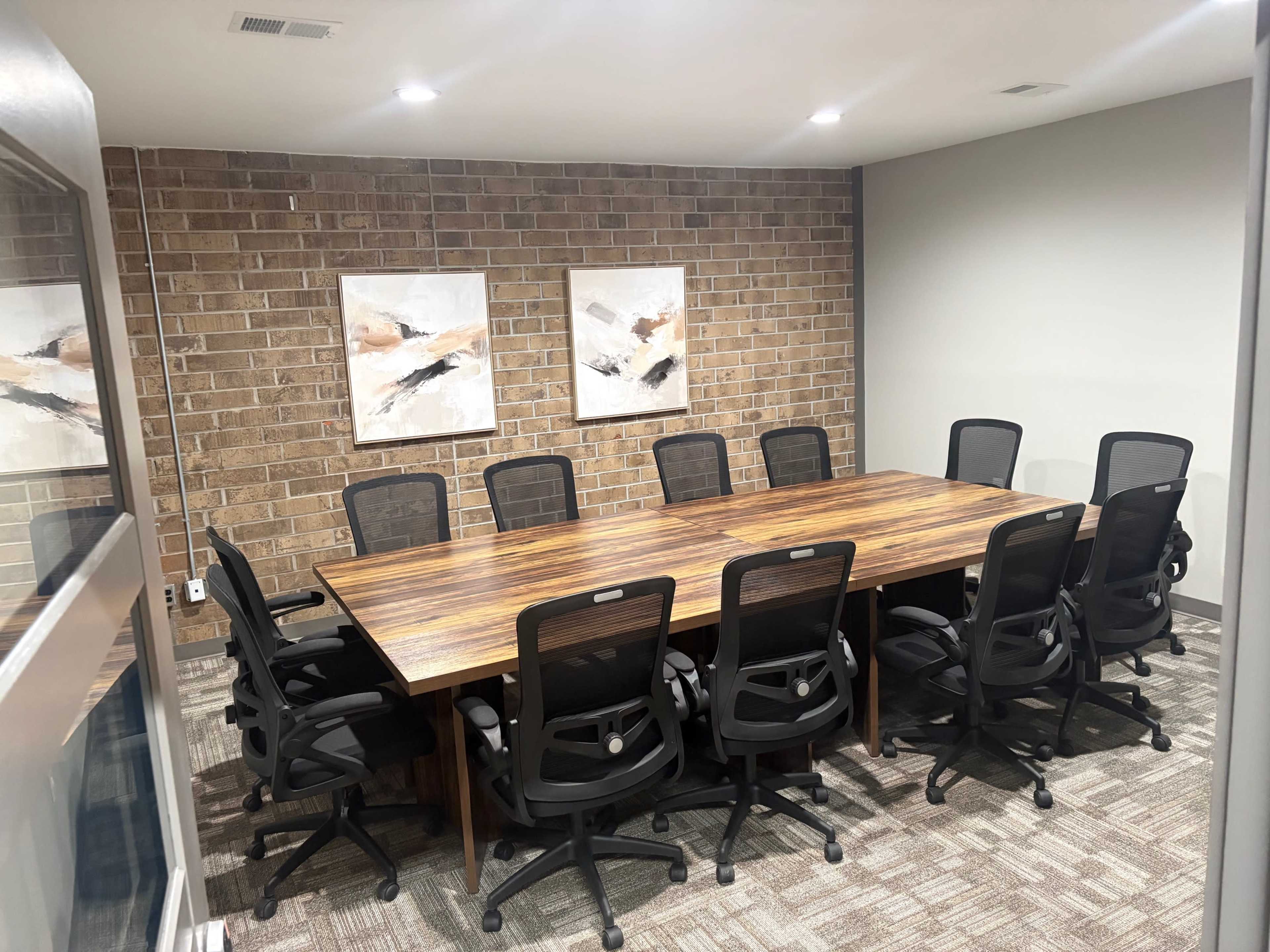 The image shows a conference room with a large wooden table surrounded by twelve black mesh chairs, set against a brick wall and two framed artworks.