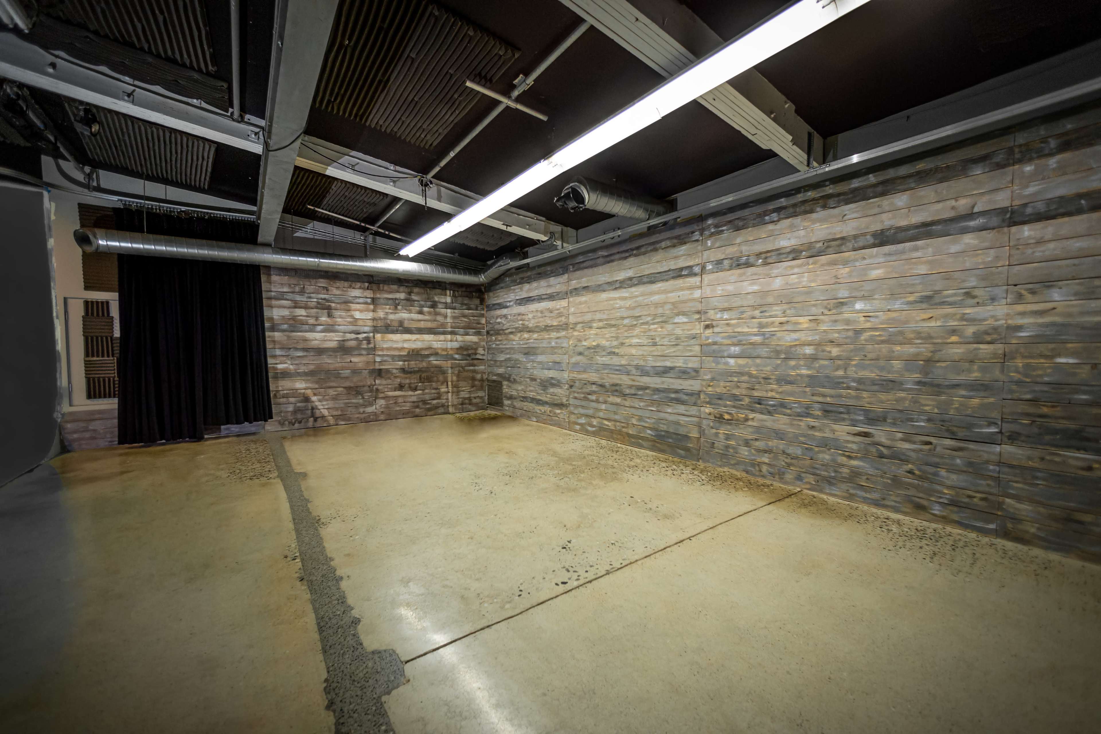 An empty room with wooden walls and a concrete floor, featuring exposed piping and overhead lighting.