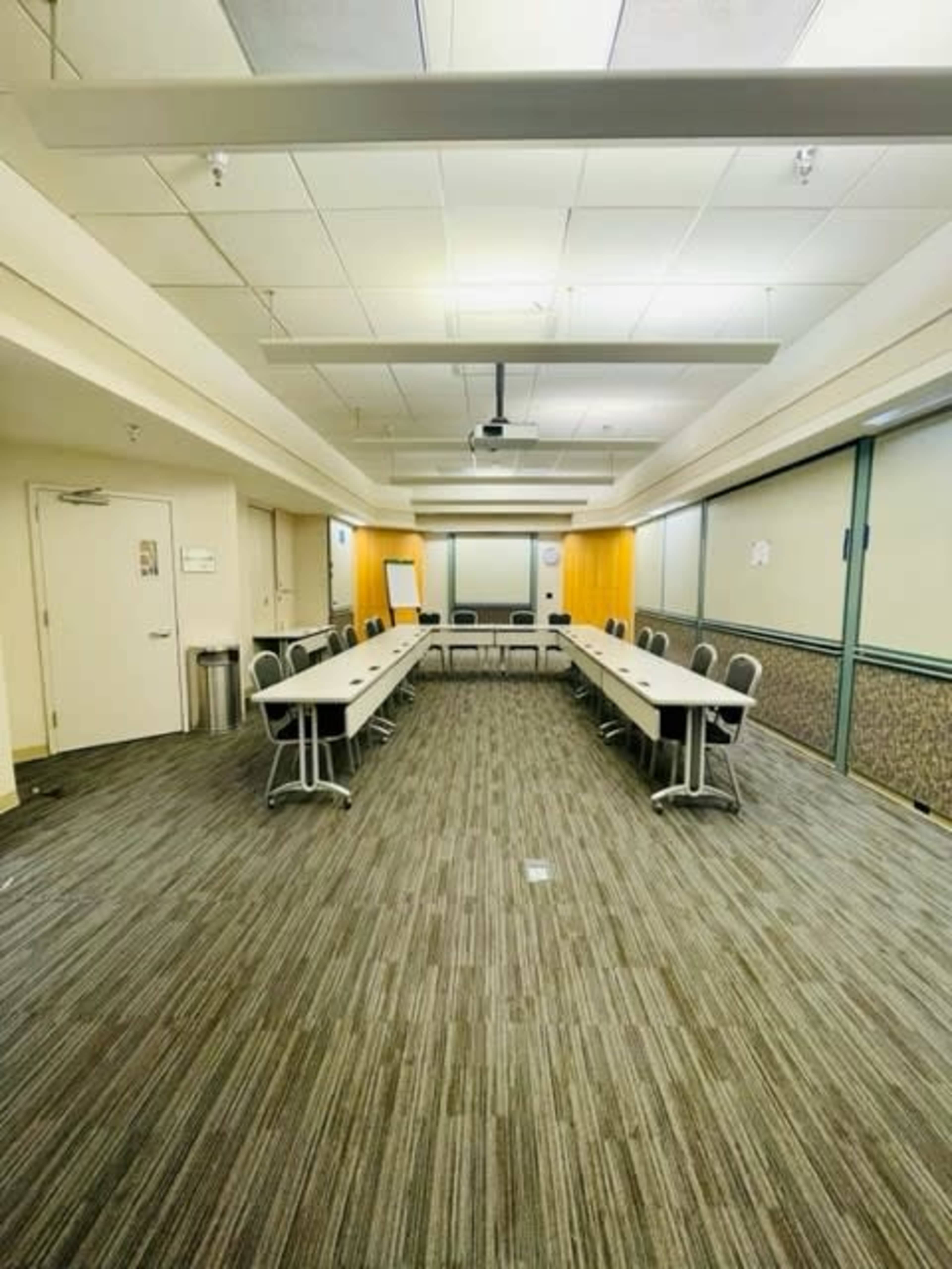 A meeting room arranged with long tables and chairs in a symmetrical layout, featuring a projector and a whiteboard at one end.