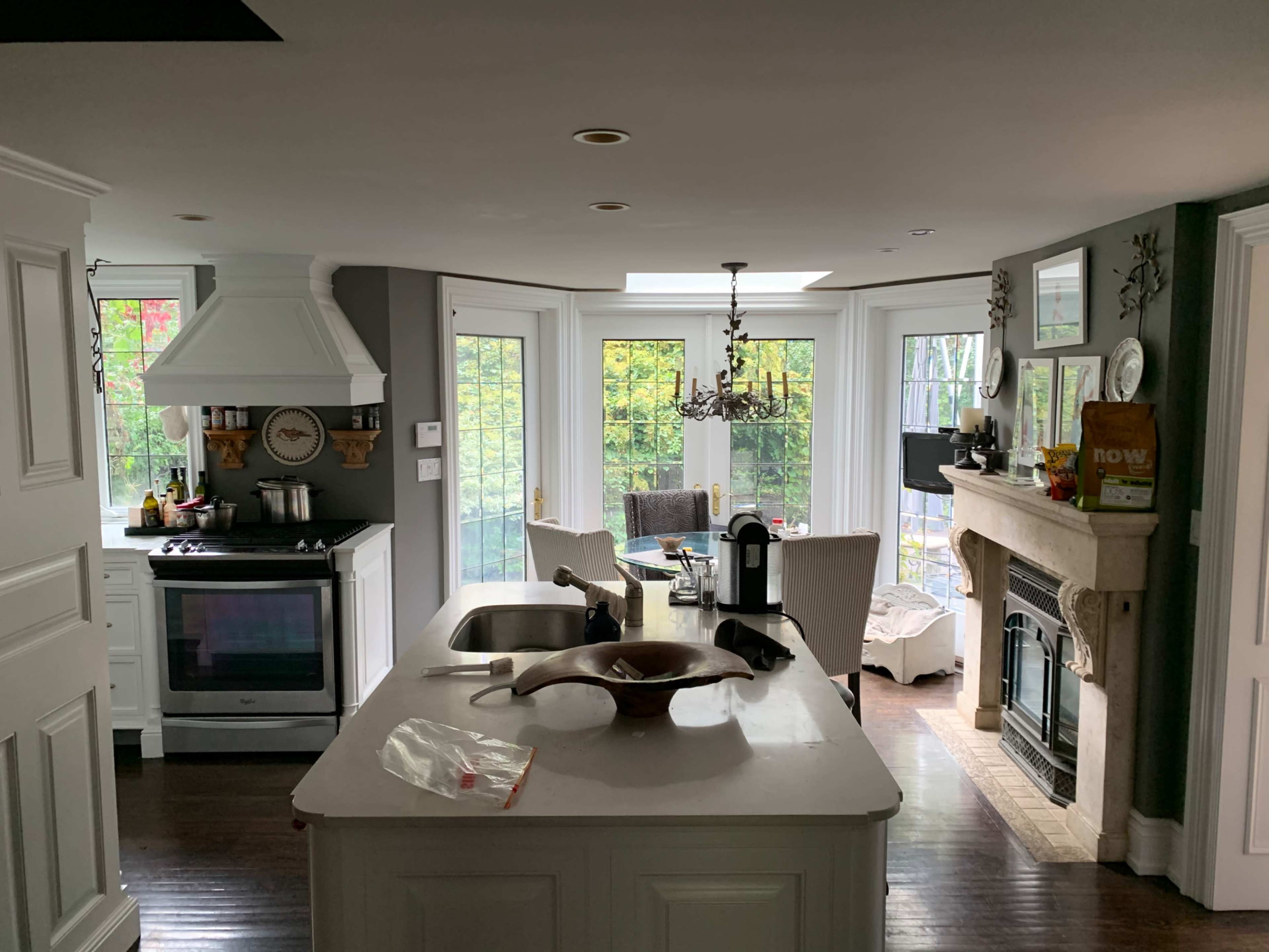A modern kitchen with white cabinetry and a central island, featuring a view of a dining area with a fireplace and large windows.
